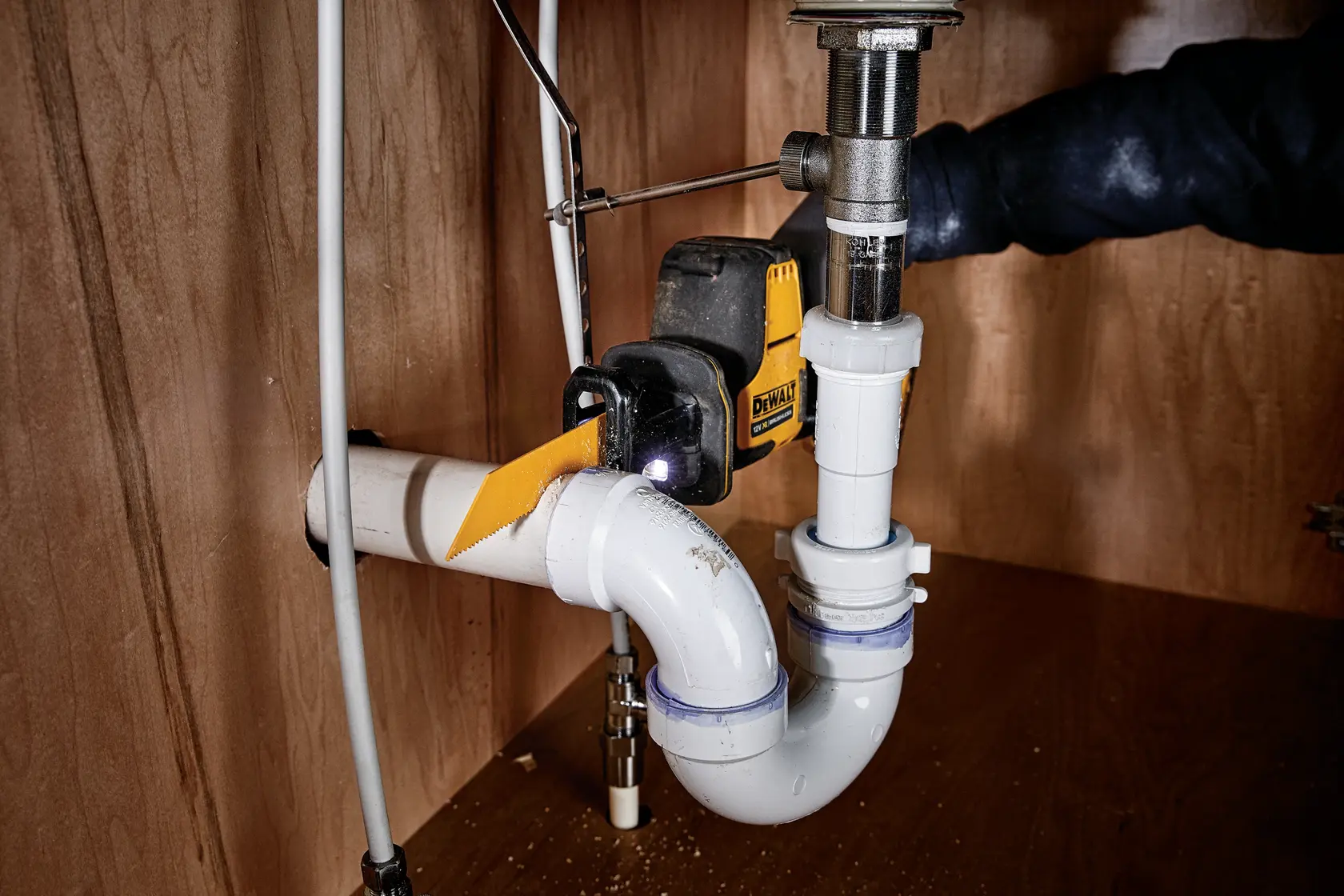 DEWALT tool cutting white PVC plumbing pipe under a sink in a wooden cabinet.