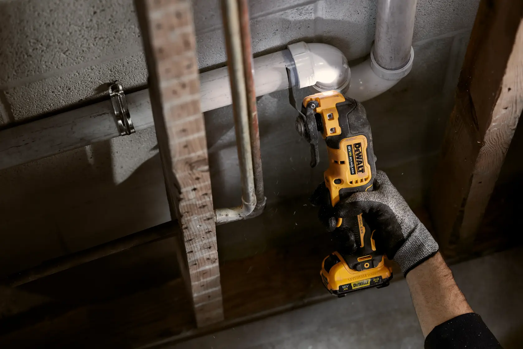 Gloved hand uses DEWALT cordless tool to cut pipe near wall and wooden beams in a construction setting.