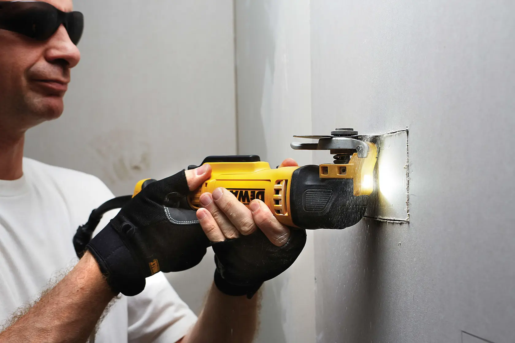 Gloved hands use a DEWALT oscillating tool to cut a square hole in a wall, tool emits light at the cut.