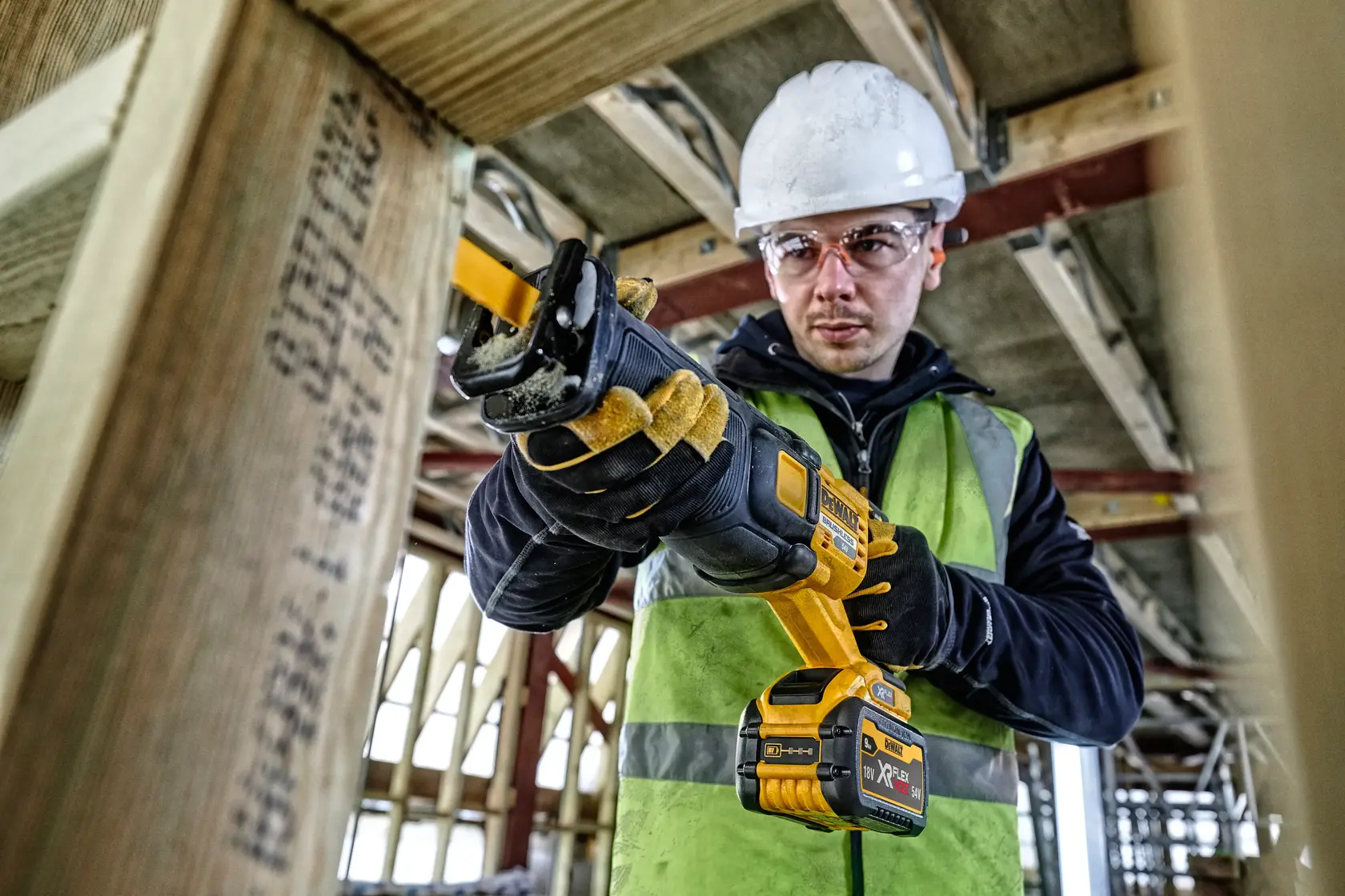 Worker with safety gear using a DEWALT cordless reciprocating saw to cut wood on a construction site.