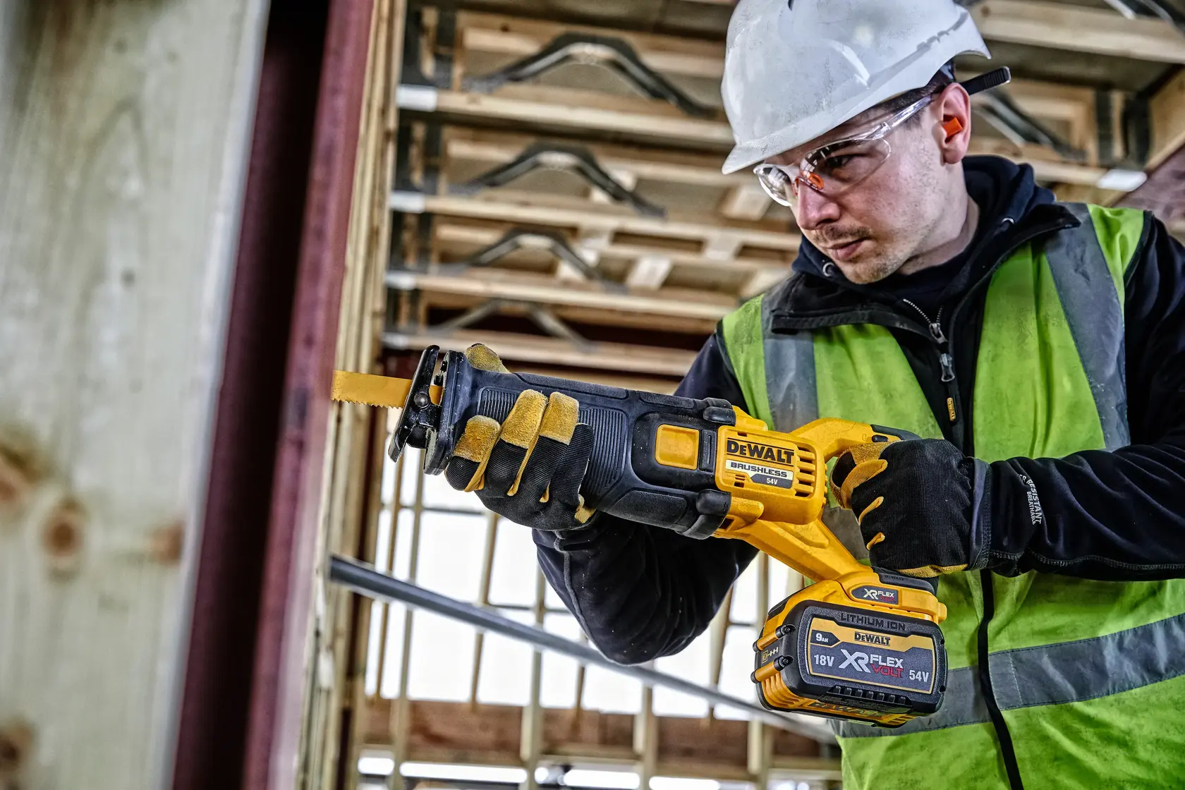 Person using a DEWALT DCS389N cordless reciprocating saw at a construction site with safety gear.
