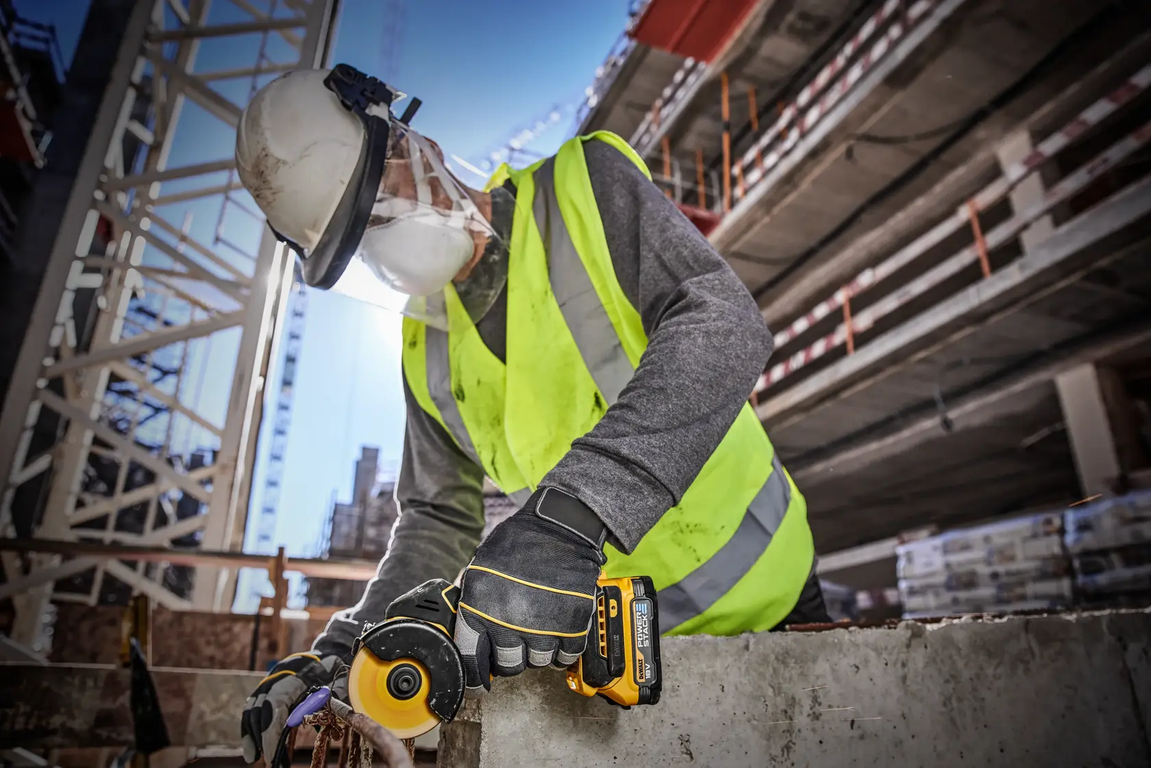 A worker wearing a hard hat, face shield, gloves, and a high-visibility vest uses a DEWALT cordless angle grinder (model DCS438E2T) on a concrete slab at a construction site.