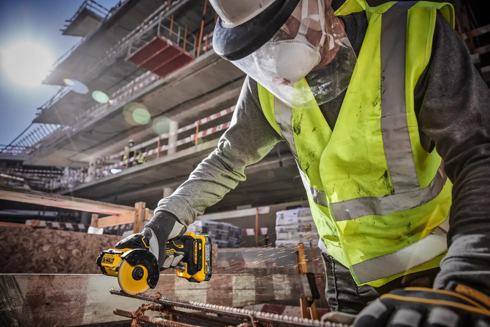 A worker wearing a high-visibility vest, hard hat, face shield, and gloves is using a DEWALT cordless rotary tool to cut metal rebar at a construction site.