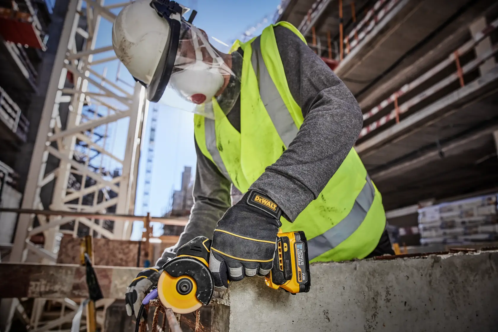 A construction worker wearing a safety helmet, face shield, reflective vest, and gloves is using a DEWALT DCS438N cordless tool to cut through concrete at an outdoor construction site.