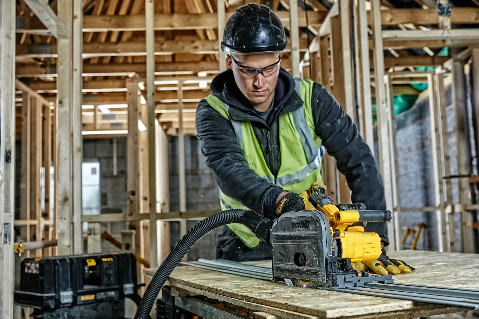 Worker uses DEWALT DCS520NT track saw to cut wood in a building under construction. Safety gear and tools are visible.