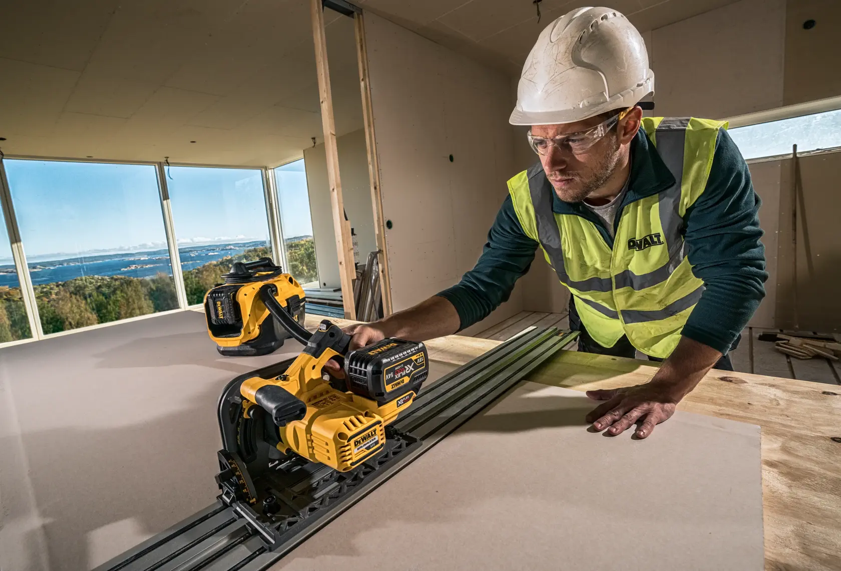 Worker using DEWALT cordless track saw DCS520NT on wood board in modern construction site with safety gear.