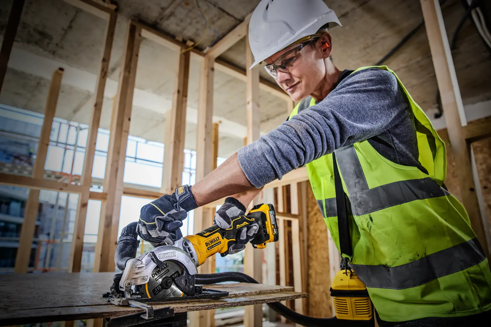 A construction worker wearing a safety vest, gloves, and a helmet uses a DEWALT cordless circular saw (model DCS571E1T) to cut a piece of wood at a building site with exposed framing.