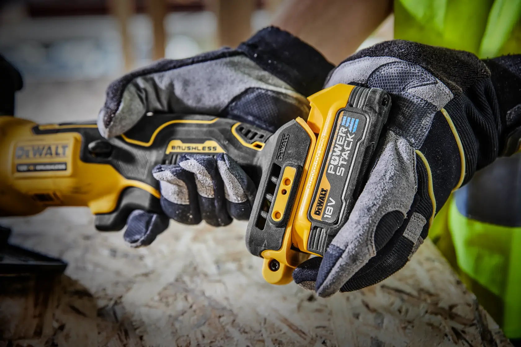 Close-up of a person wearing work gloves attaching an 18V DEWALT POWERSTACK battery to a DEWALT brushless power tool on a wooden surface.