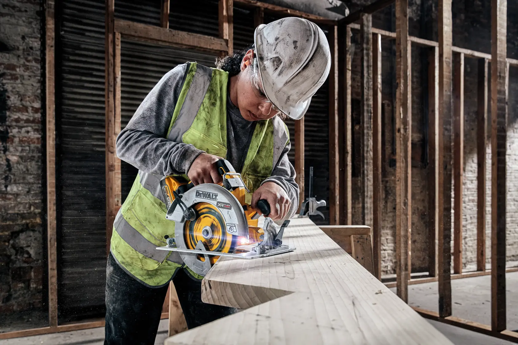 A person wearing a safety vest and hard hat using a DEWALT DCS573H2T cordless circular saw to cut a large wooden plank in an unfinished construction site.