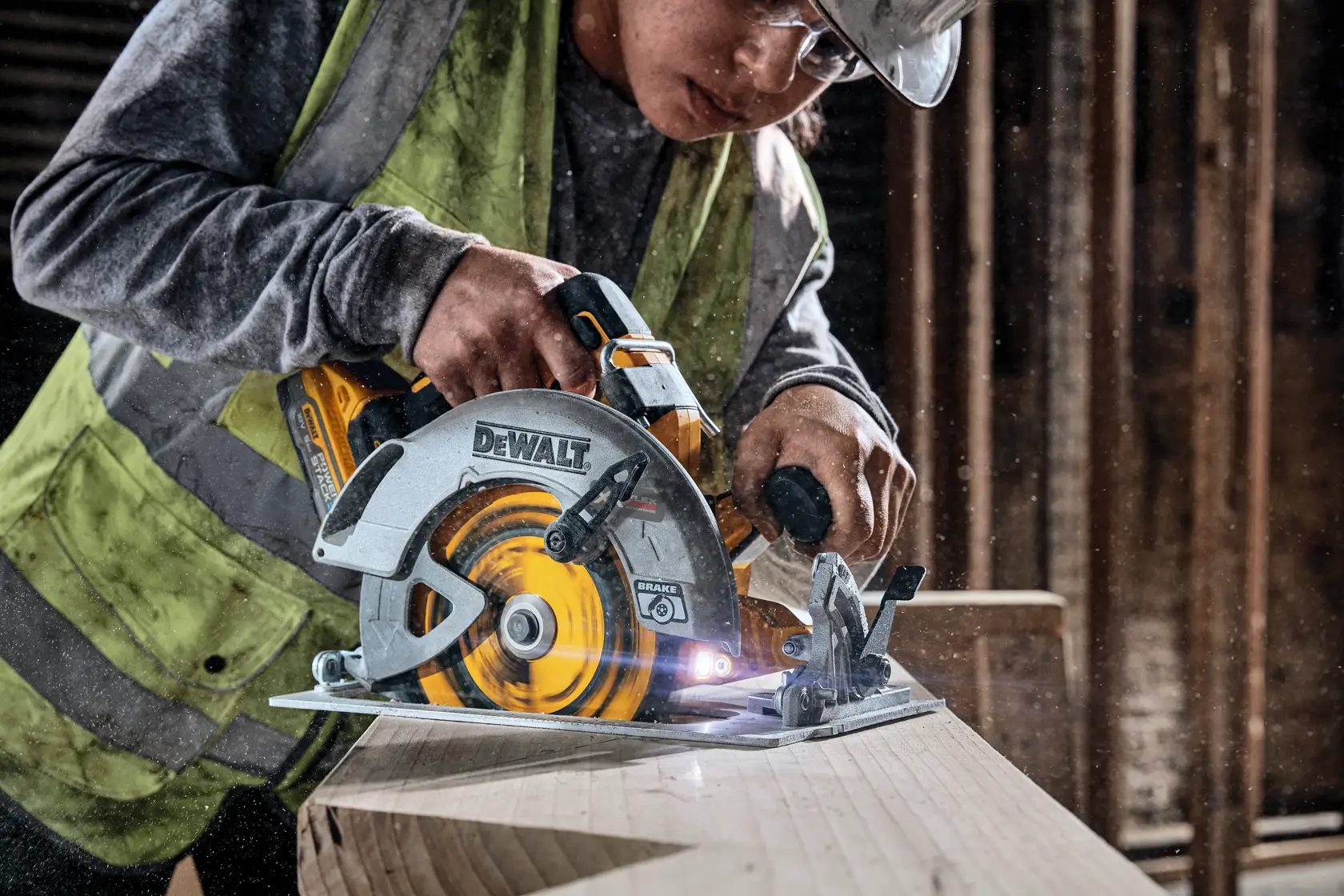A person wearing safety gear is using a DEWALT cordless circular saw to cut a wooden beam in a workshop or construction site.