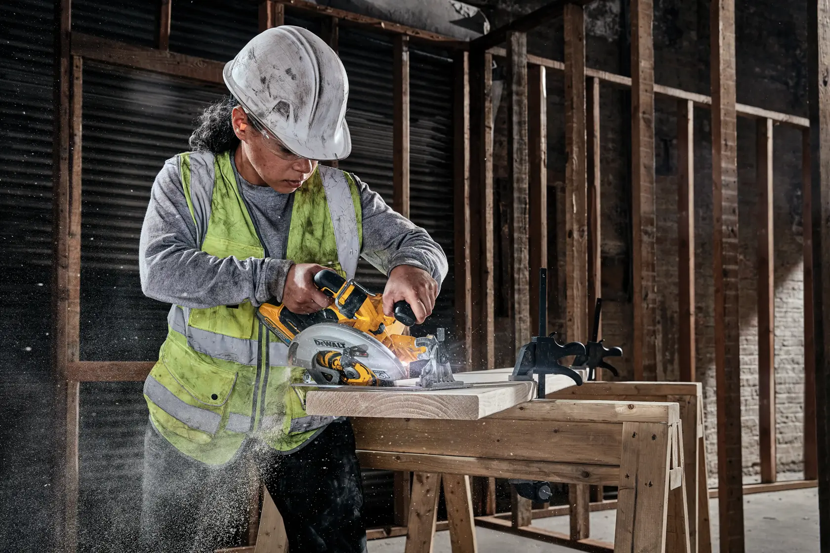 A construction worker wearing a safety helmet and high-visibility vest is using a DEWALT cordless circular saw to cut wooden planks on a workbench in an unfinished building.