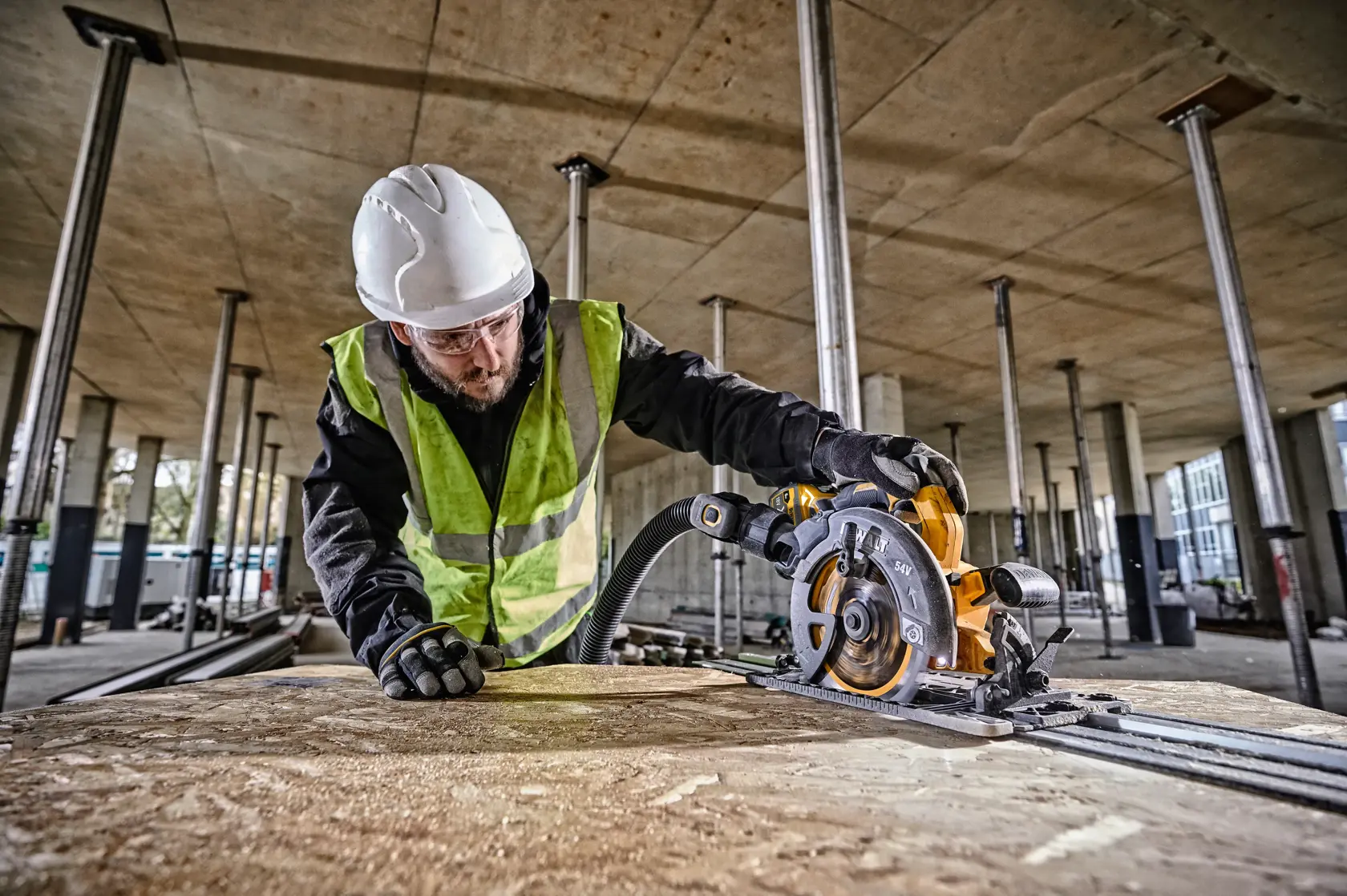 Worker using DEWALT DCS579T2 circular saw on wood panel at a construction site, wearing safety gear.