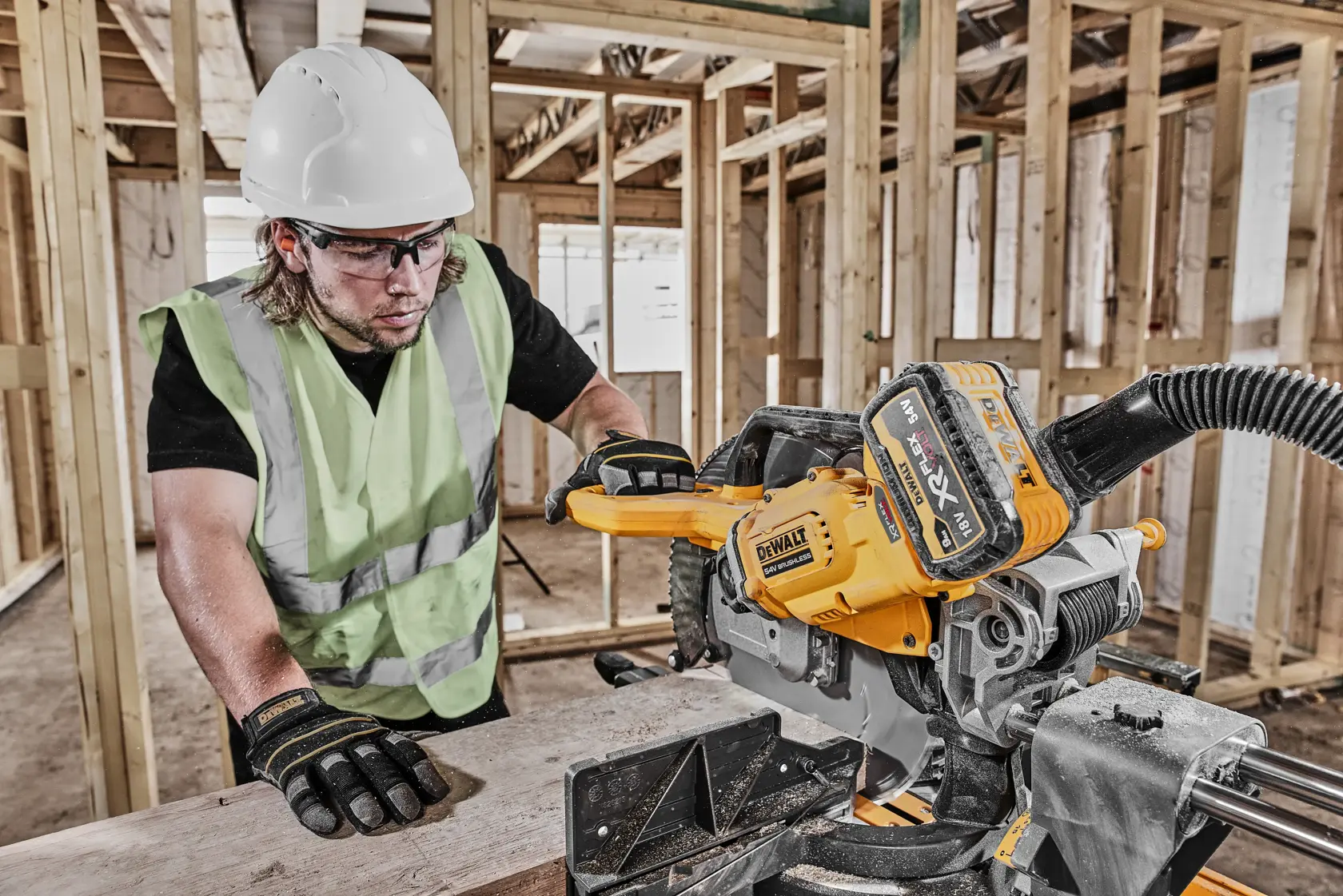 Worker using DEWALT DCS781N saw on a wooden plank at a construction site, wearing safety gear.