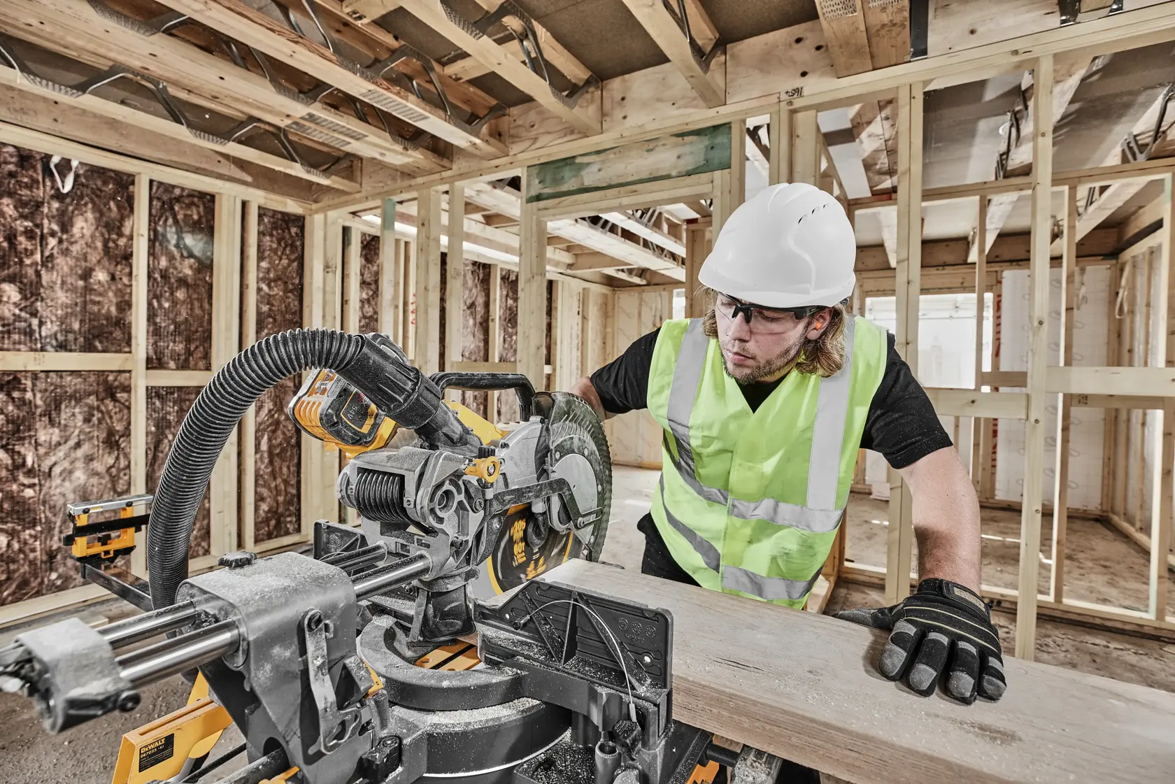 Worker uses DEWALT miter saw DCS781N on wood in a construction site wearing safety gear.