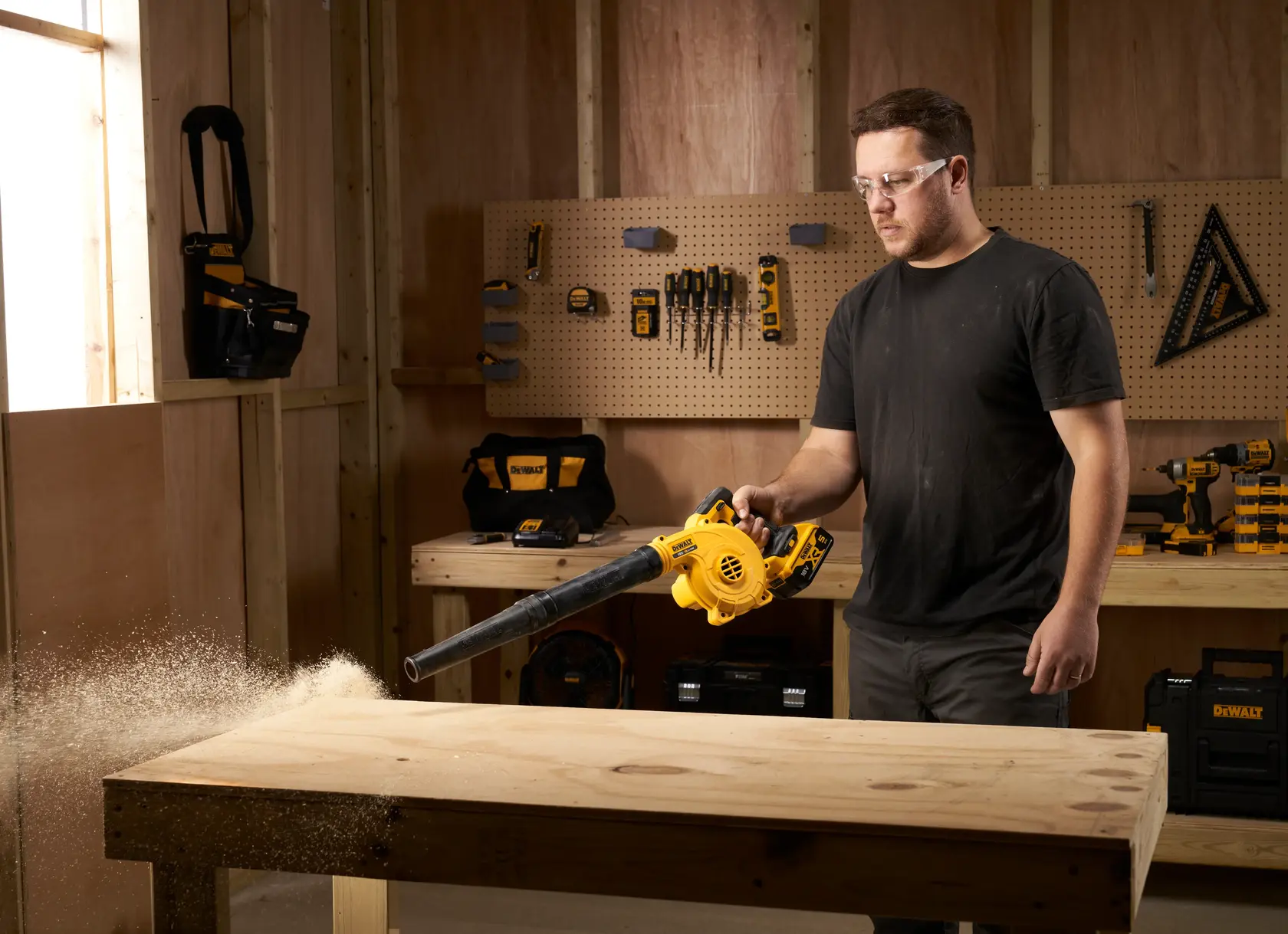 Person using a DEWALT cordless blower to clean sawdust from a wooden workbench in a workshop.