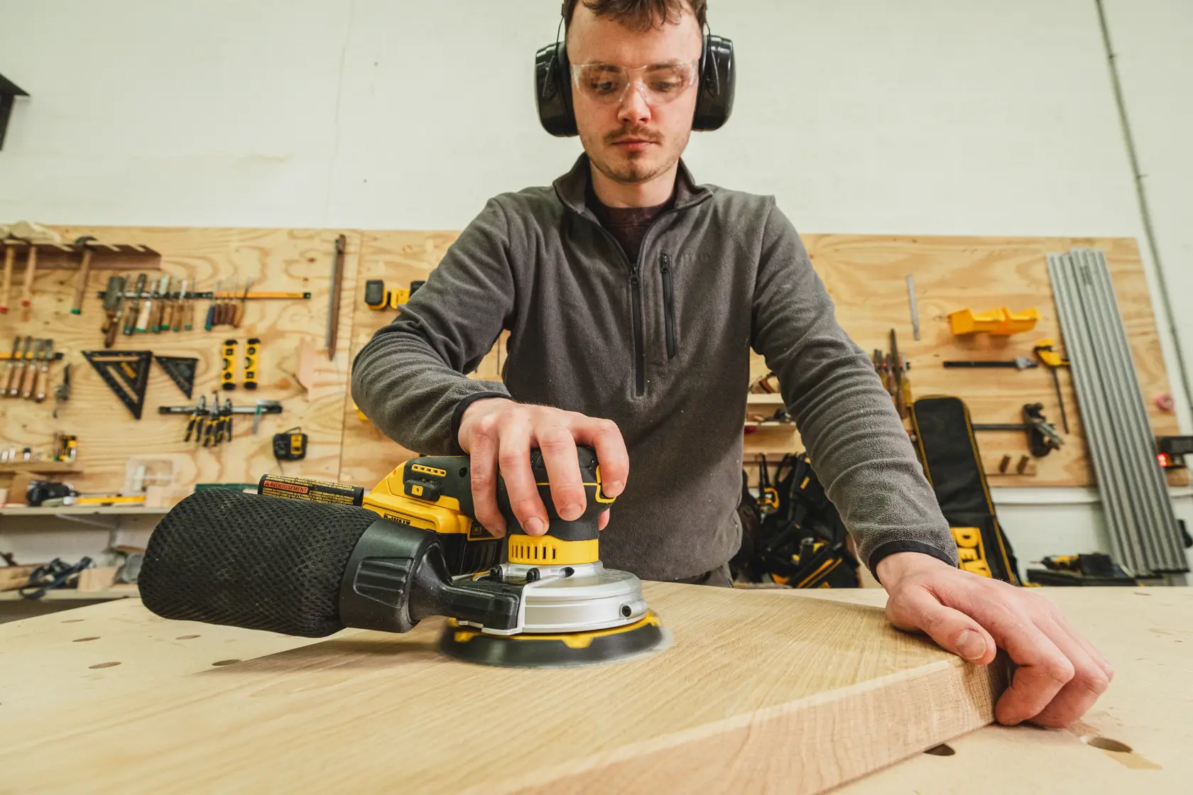 Person using a DEWALT sander on a wooden board in a workshop with tools in the background.