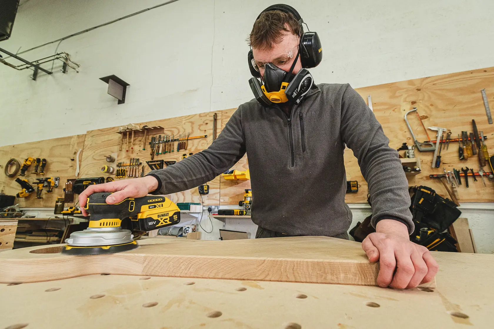 Person sanding wood with a DEWALT sander, wearing safety gear in a workshop with tools on the wall.