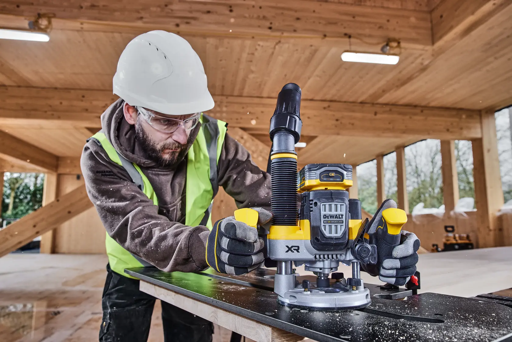 Worker uses DEWALT router DCW620NT on wood in construction site, wearing safety gear and gloves.