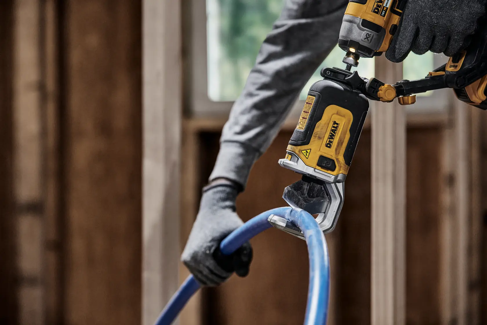 Gloved hand uses DEWALT tool to cut a blue pipe in a construction setting with wooden framing.