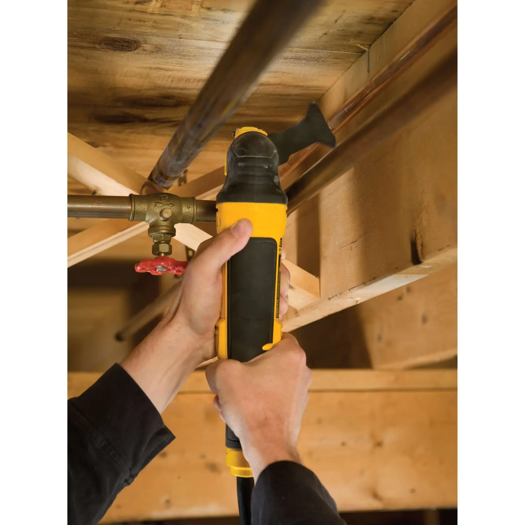 Hands using a DEWALT power tool to cut a metal pipe under a wooden ceiling structure.