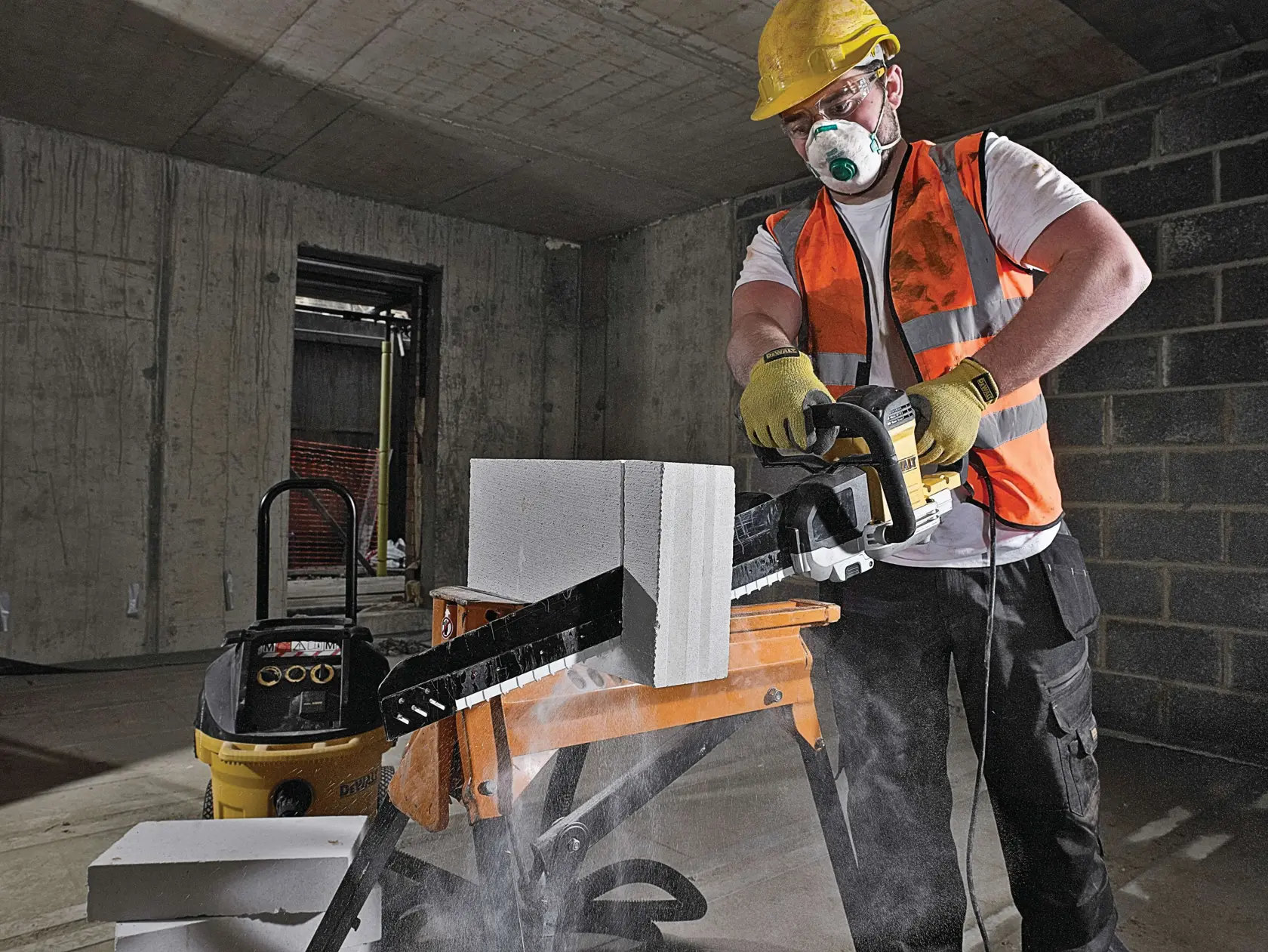 Worker uses DEWALT power tool to cut concrete block indoors, wearing safety vest, gloves, and helmet.