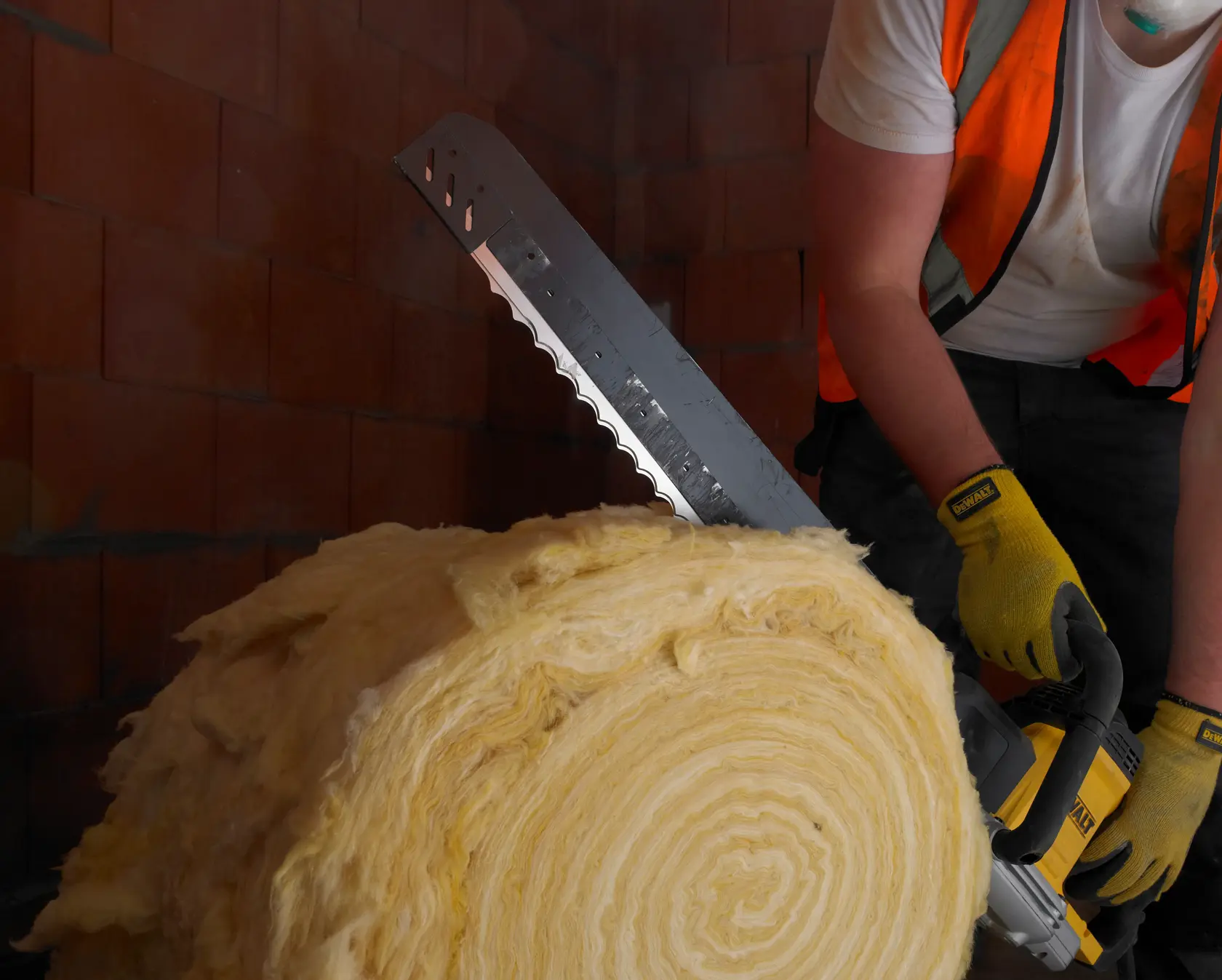 Person cuts insulation roll with a DEWALT saw, wearing gloves and safety vest in a brick-walled area.