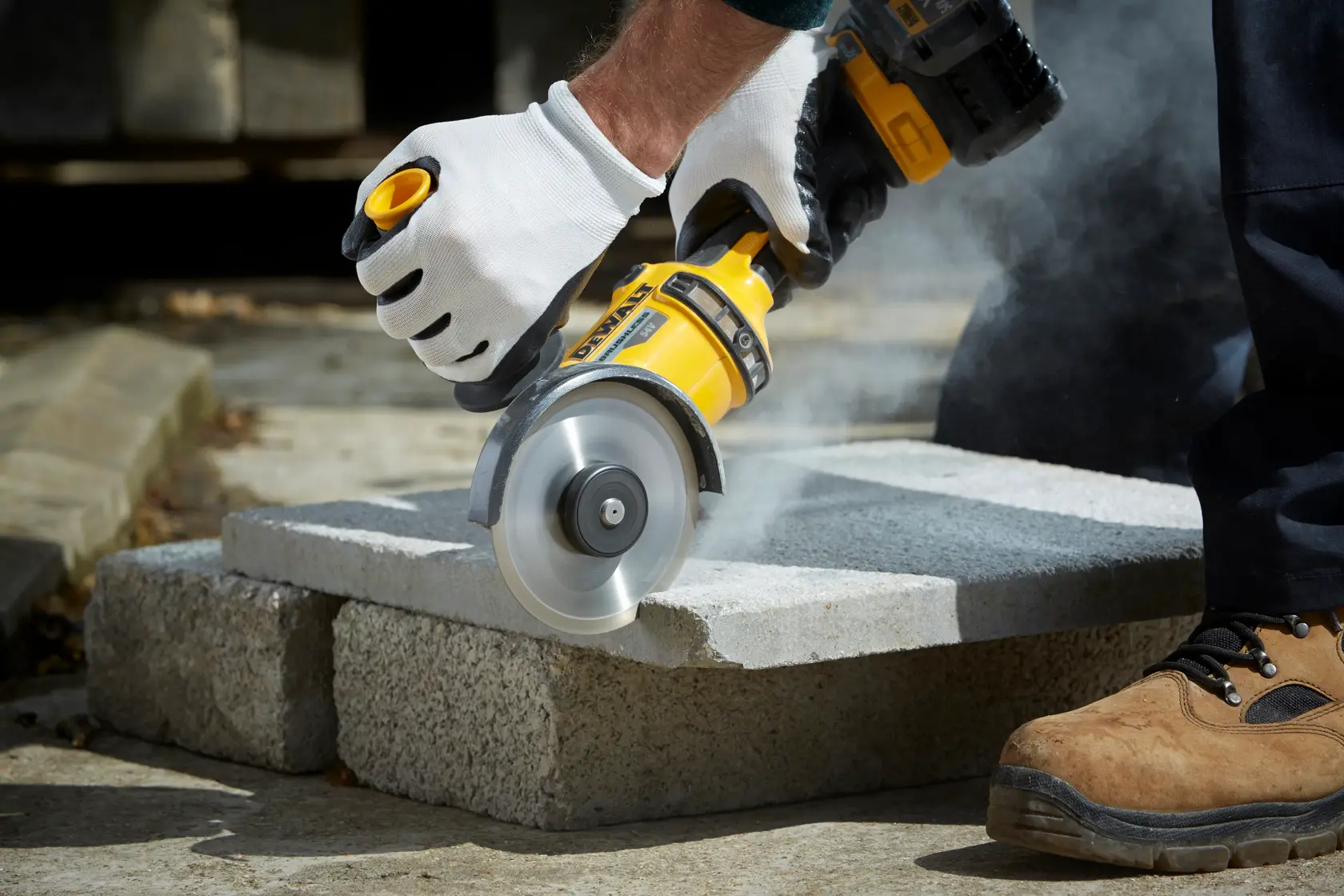 Person uses DEWALT angle grinder DT3711 to cut concrete block outdoors, wearing gloves and safety shoes.