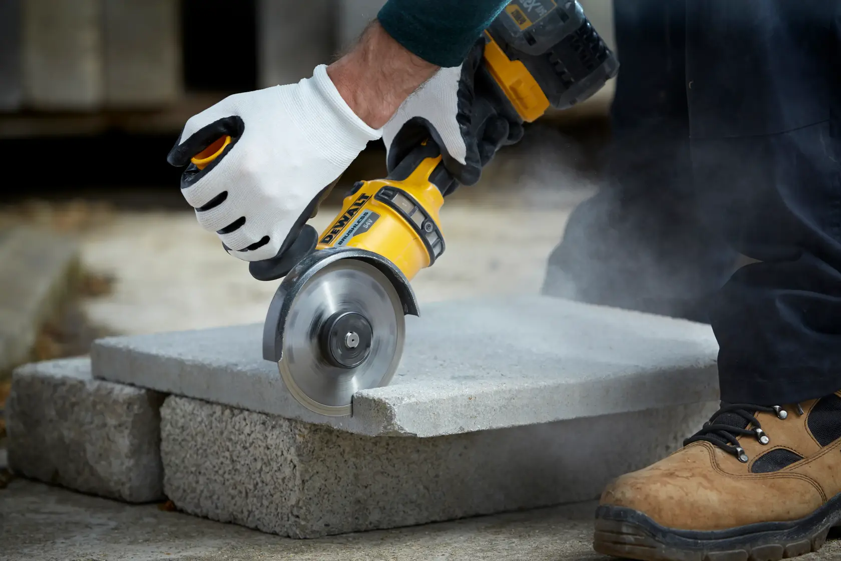 A gloved hand uses a DEWALT tool to cut a concrete slab, with dust rising during the process.