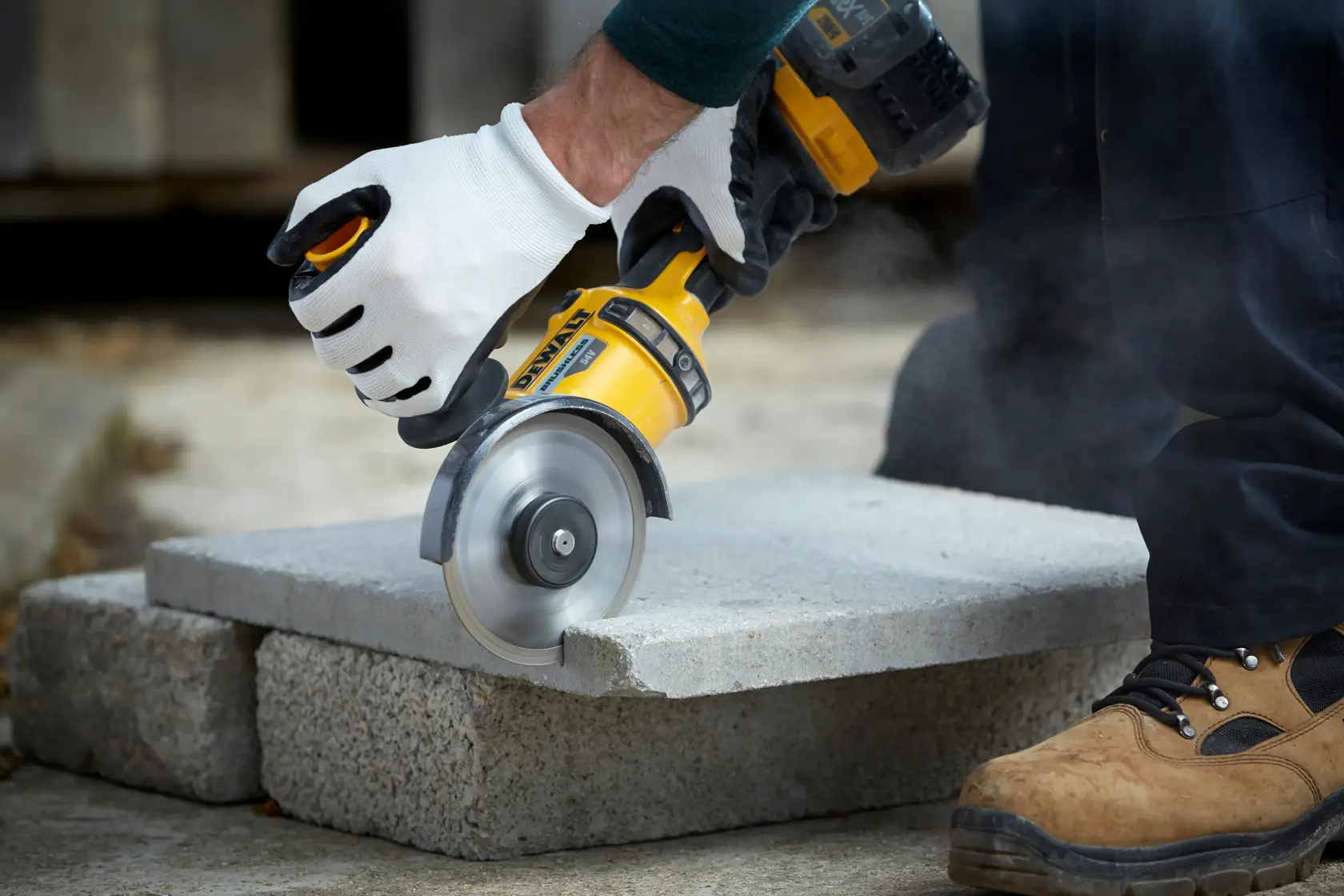 Person cuts concrete block with a DEWALT power tool, wearing gloves and work boots outdoors.
