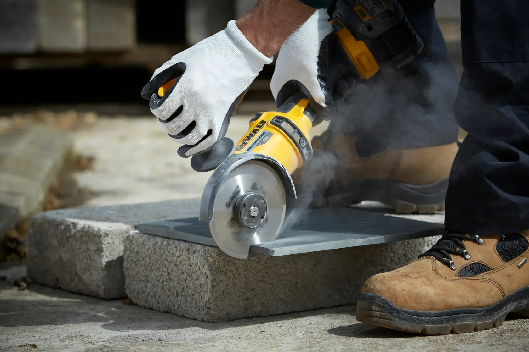 Gloved hands use a DEWALT angle grinder to cut stone outdoors, generating dust.