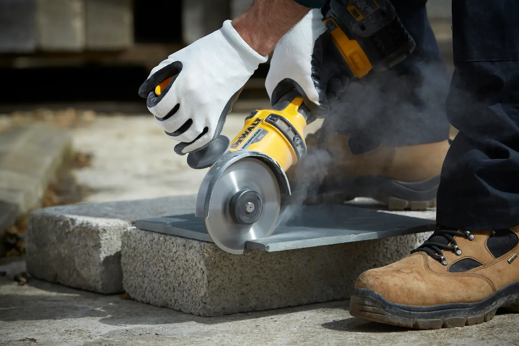 Person using a DEWALT angle grinder to cut stone outdoors, wearing gloves and safety boots.