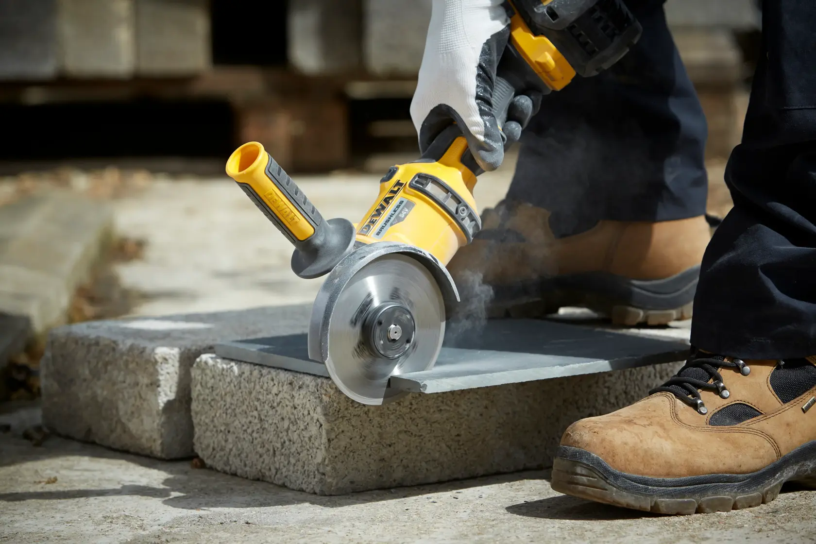Person using DEWALT angle grinder to cut a stone slab outdoors, wearing gloves and safety footwear.