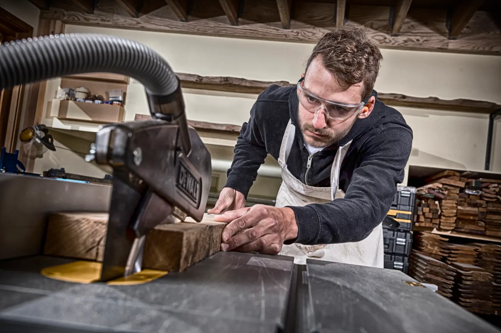 Person cutting wood with DEWALT saw in workshop, wearing apron and focused on woodworking task.