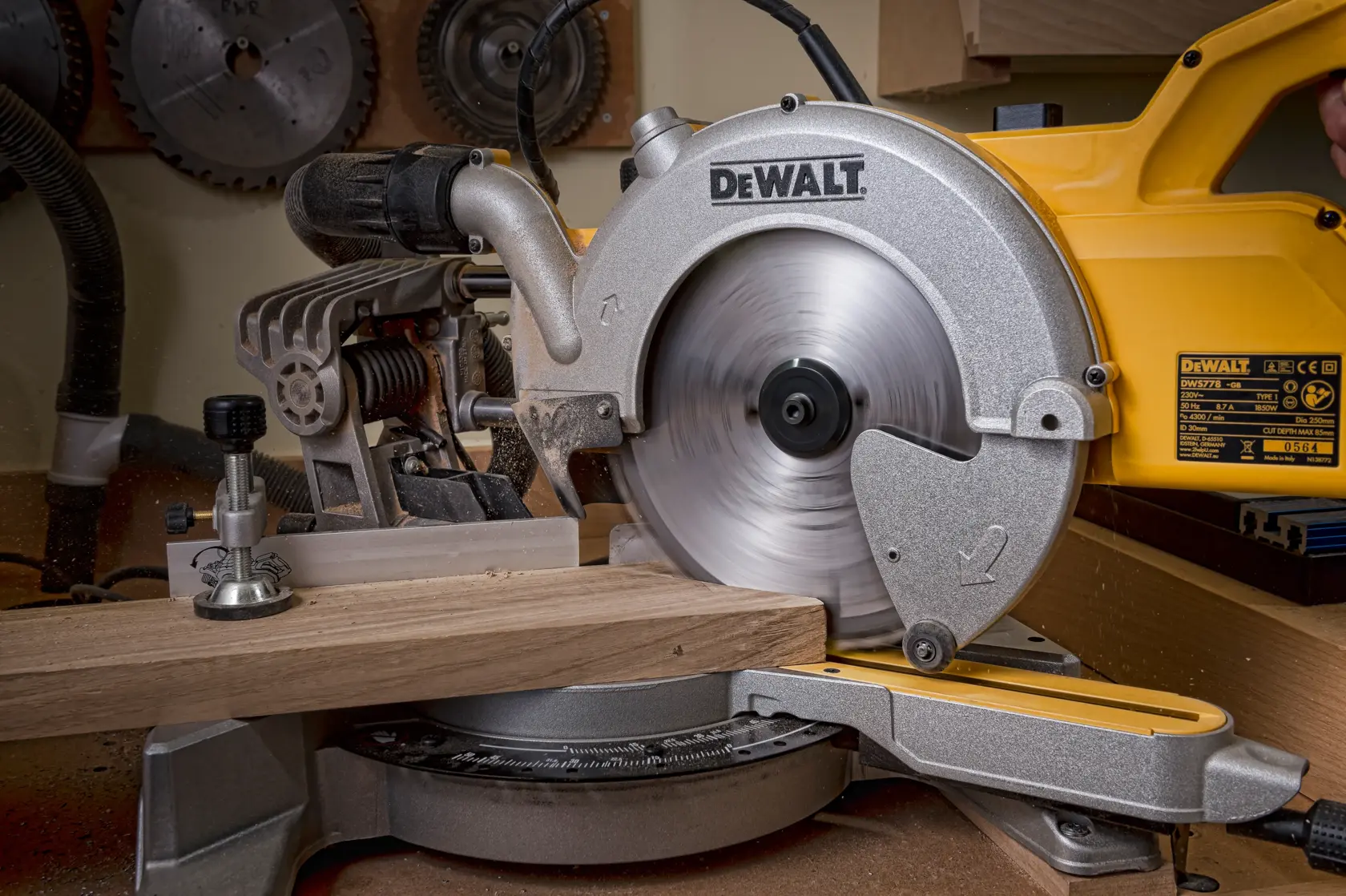DEWALT miter saw cutting wood plank in a workshop with saw blades on the wall in the background.