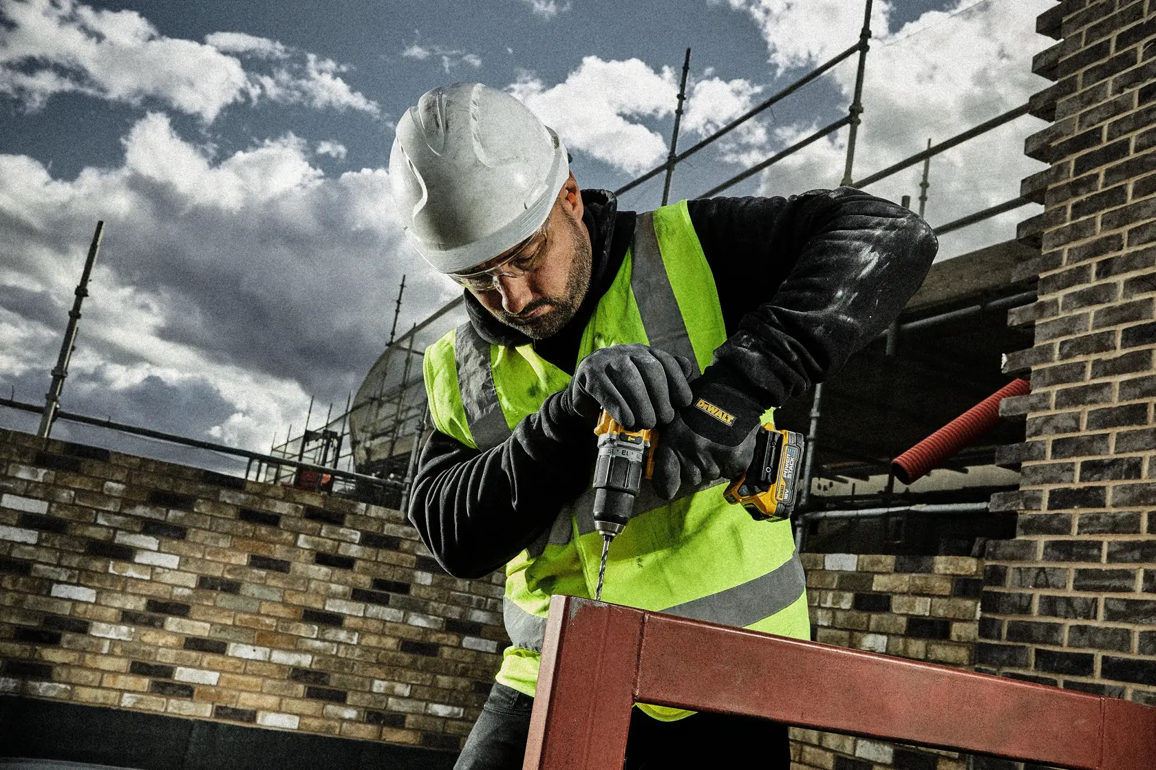 Worker in safety gear uses a DEWALT drill outdoors on a metal frame at a construction site.