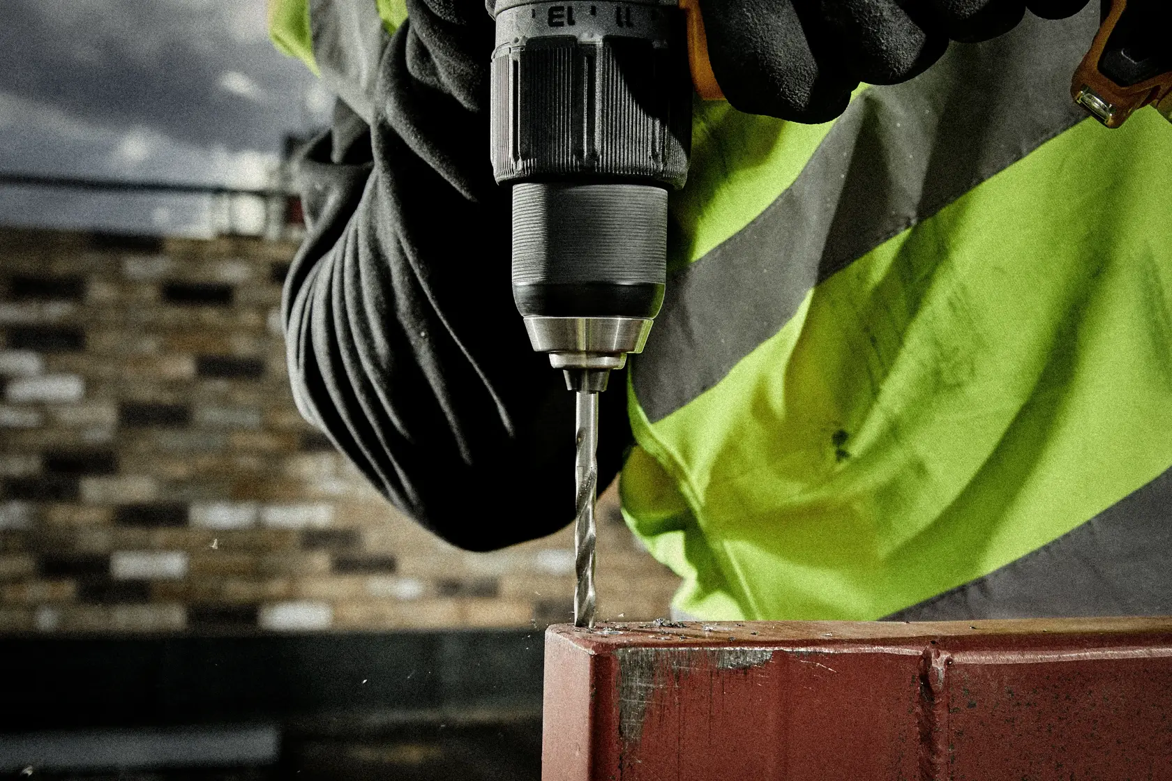 Close-up of gloved hand using a DEWALT drill to drill into metal, person in high-visibility jacket.