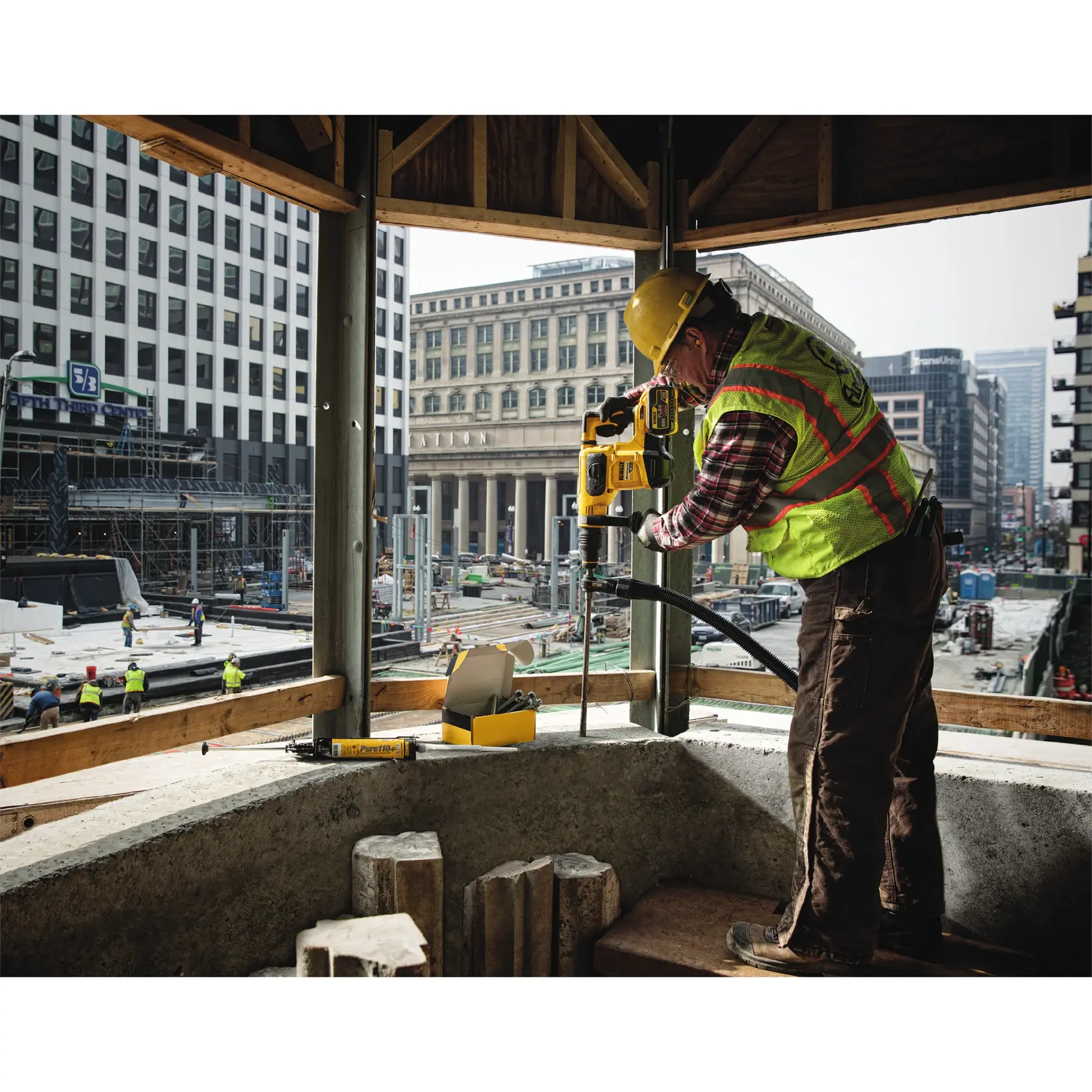 Worker uses DEWALT drill at construction site in city, wearing safety gear and helmet.