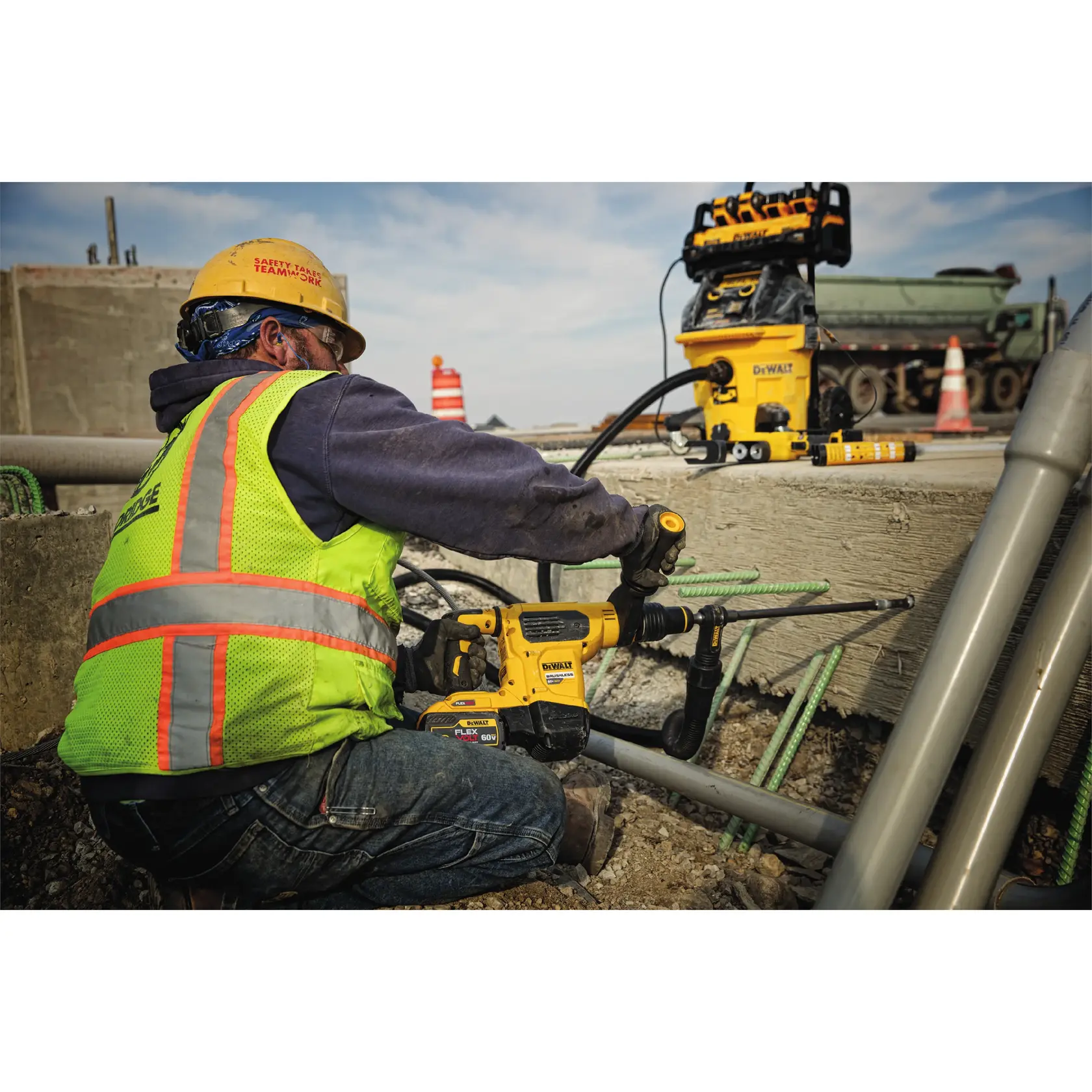 Worker in safety gear uses a DEWALT drill on concrete at a construction site with tools and pipes nearby.