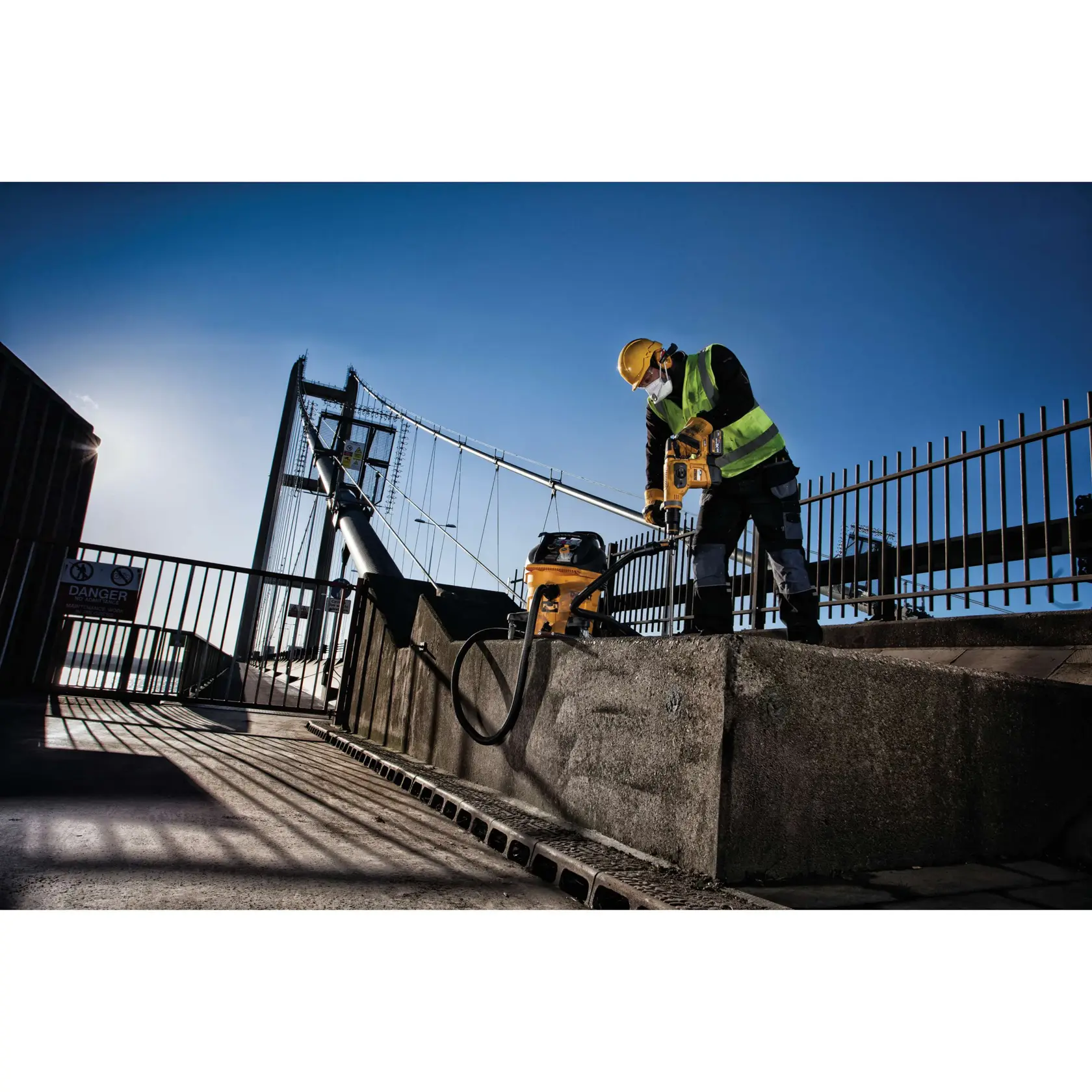 Worker uses DEWALT power tool and vacuum on concrete near a bridge, wearing safety gear and high-visibility vest.