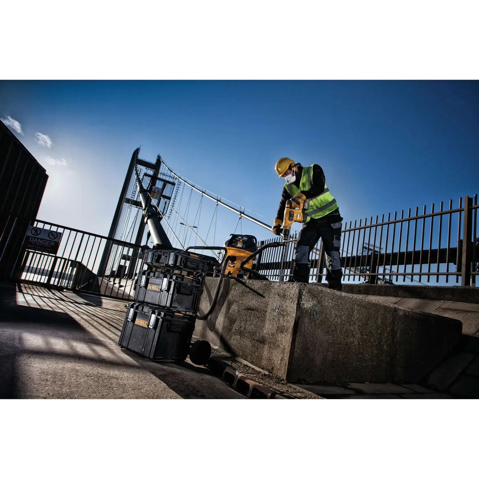Construction worker using a DEWALT tool near stacked toolboxes on an outdoor site with a bridge in the background.