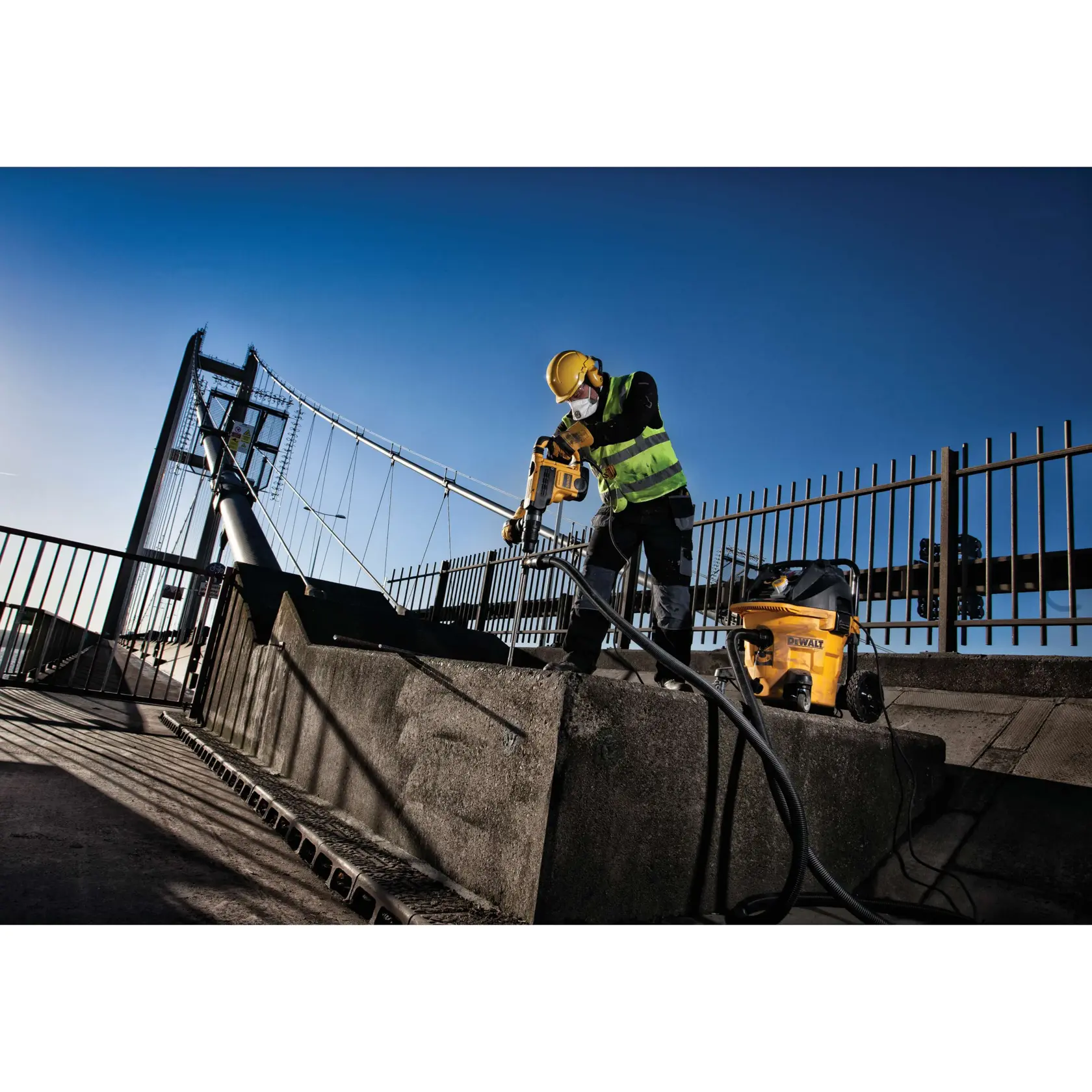 Worker uses DEWALT drill on concrete outdoors by a bridge, wearing safety gear and high-visibility vest.
