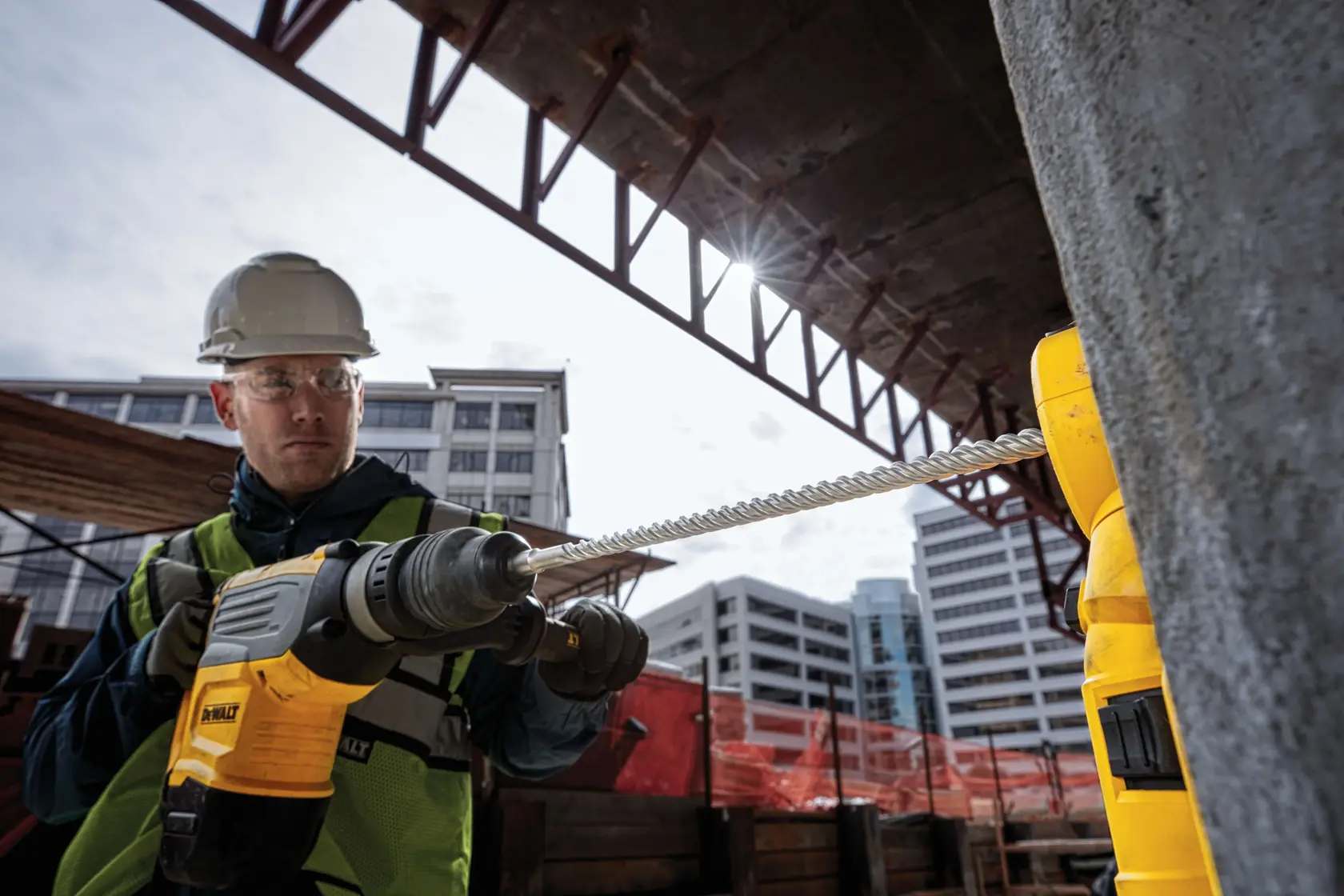 Worker uses a DEWALT drill on a construction site with buildings in the background.