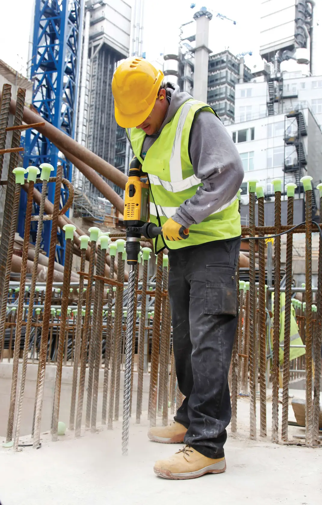 Worker using a DEWALT drill at construction site. Wearing safety gear and surrounded by rebar and buildings.