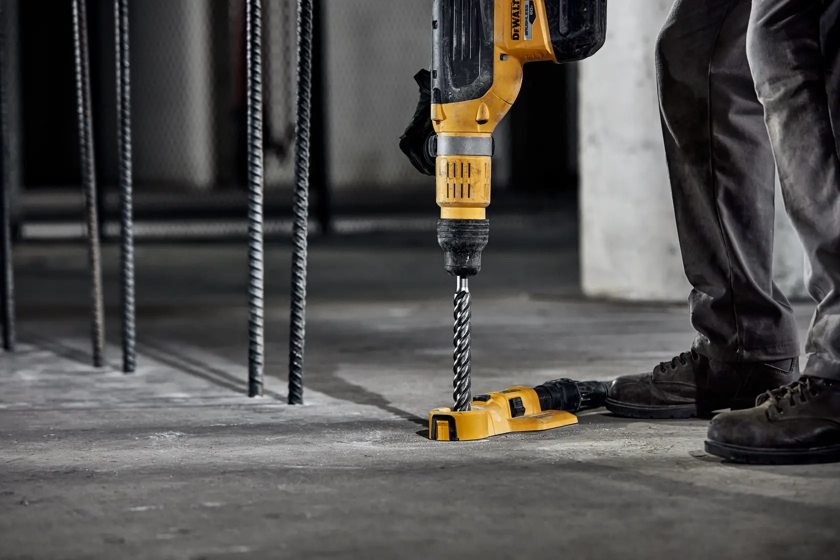 Person using DEWALT hammer drill with drill bit on concrete floor at construction site.