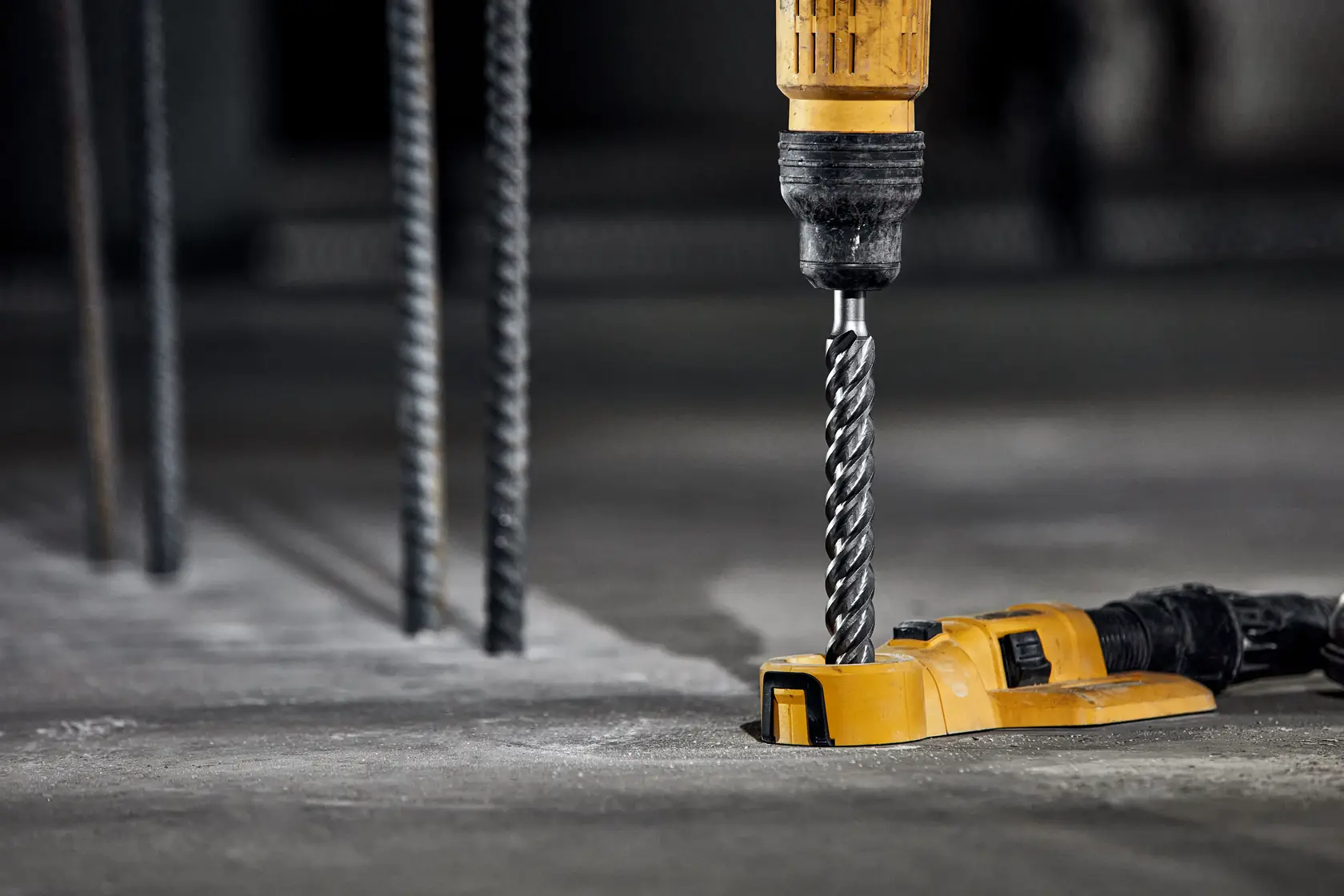 A yellow DEWALT drill with a spiral drill bit is being used to drill into a concrete surface. Metal rods are visible in the background, indicating a construction environment.