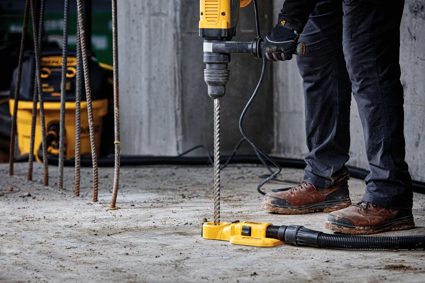 Person using a DEWALT drill with a vacuum attachment on a concrete floor at a construction site.