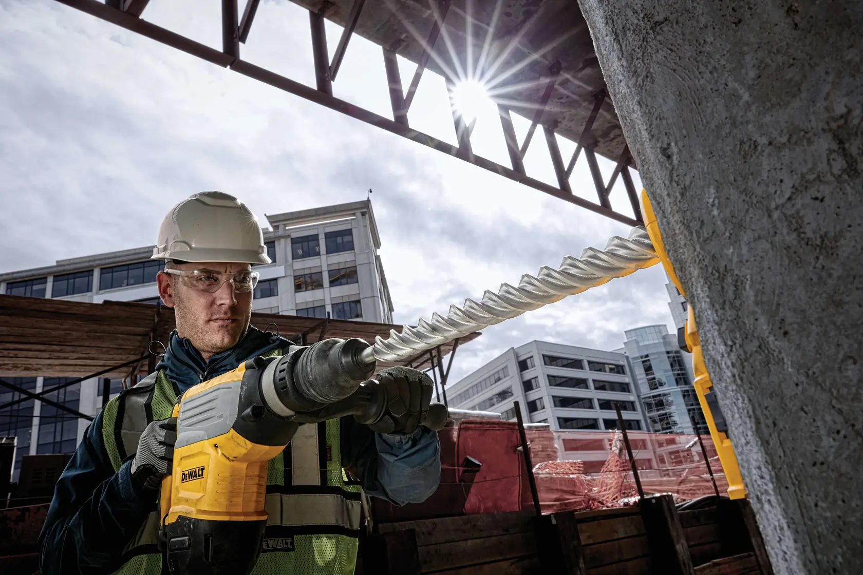 Worker uses a DEWALT hammer drill with a spiral bit on concrete at a construction site with modern buildings in background.