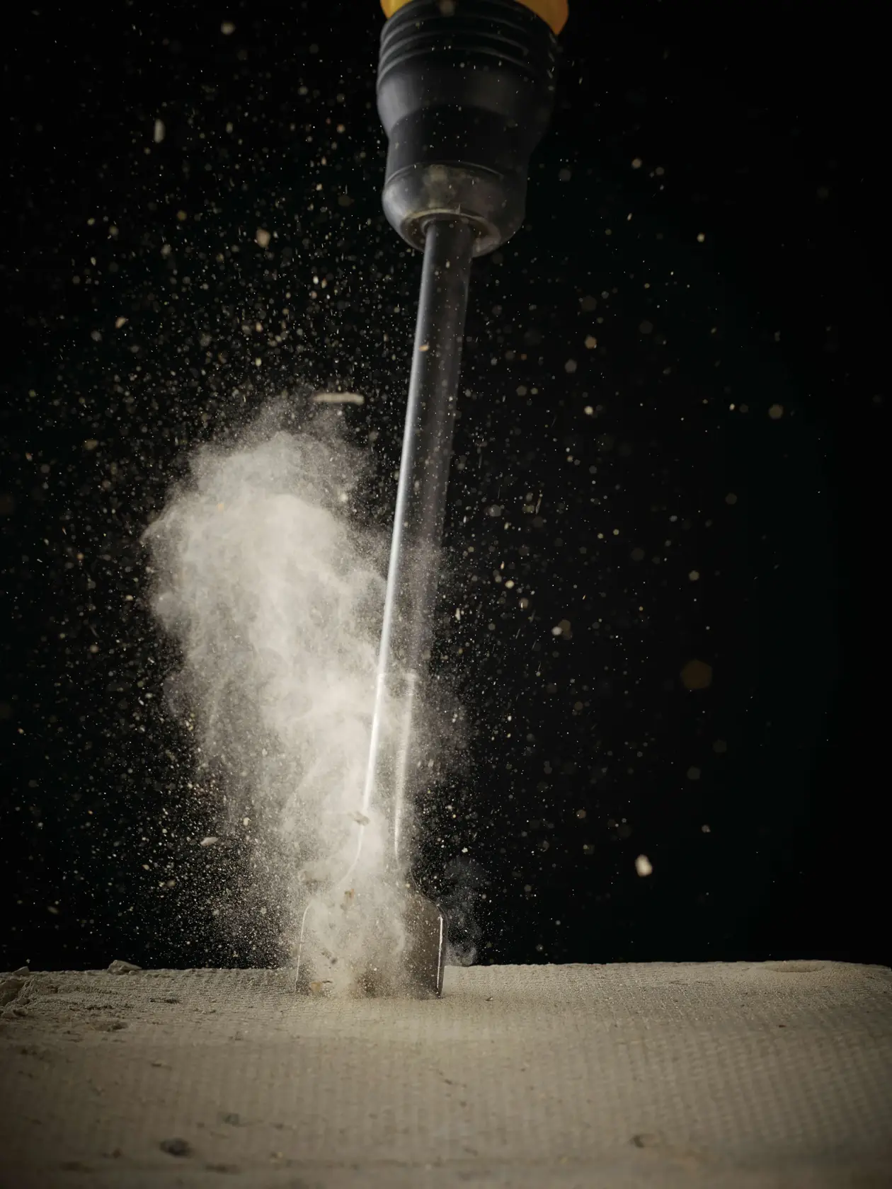 Chisel striking concrete, dust scattering on impact, close-up against dark background.