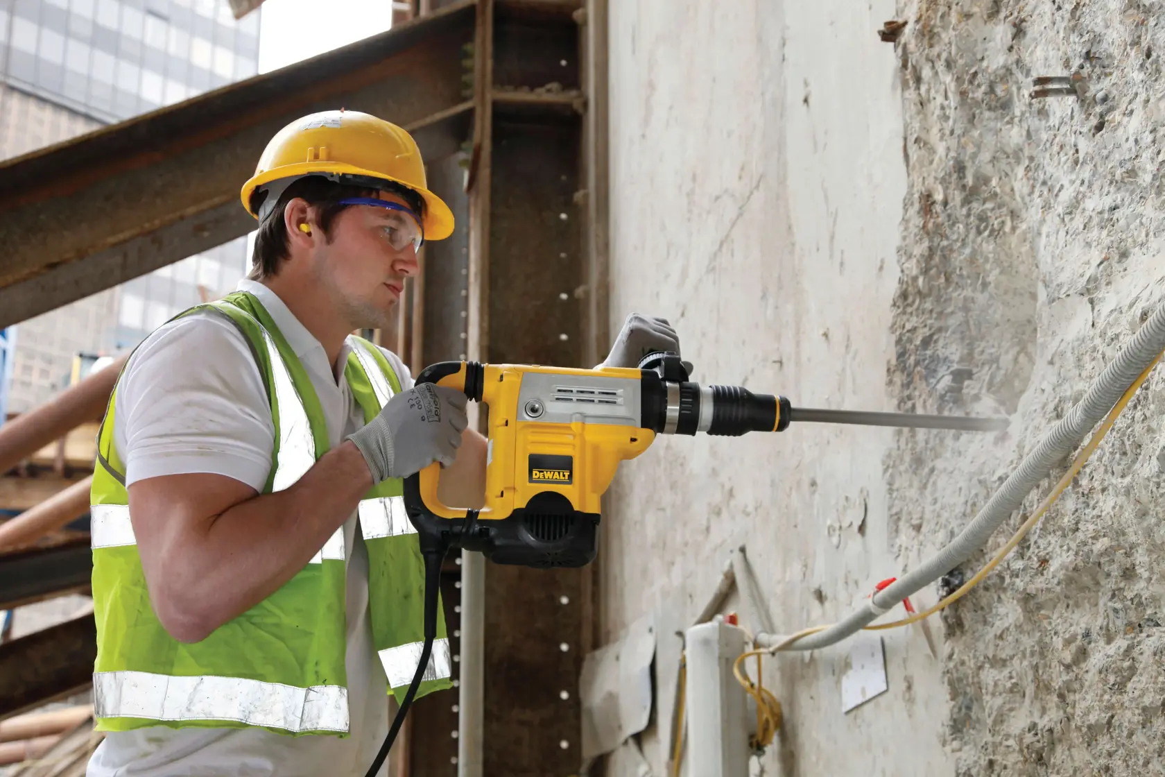 Worker uses DEWALT DT6825 chisel tool to break concrete wall at construction site, wearing safety gear.