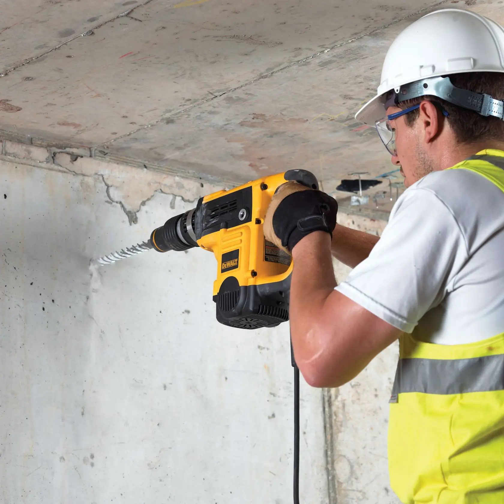 Worker drills concrete wall using a yellow DEWALT tool with safety gear and gloves.